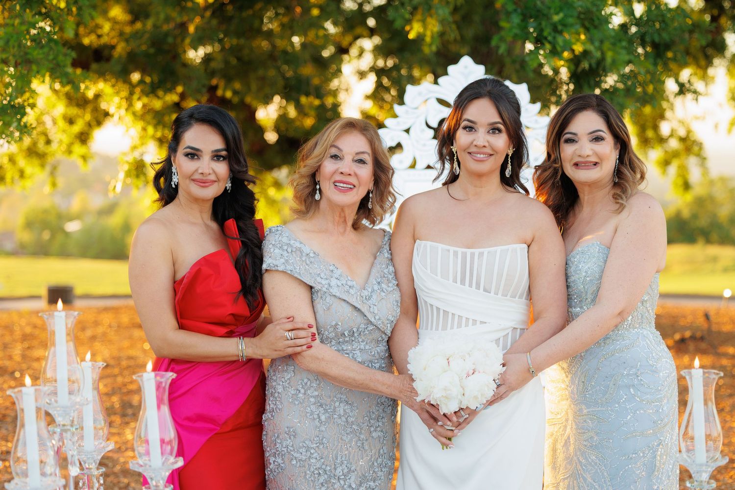 A bridal party in sparkly gowns poses together at sunset in a garden setting with golden light filtering through trees.