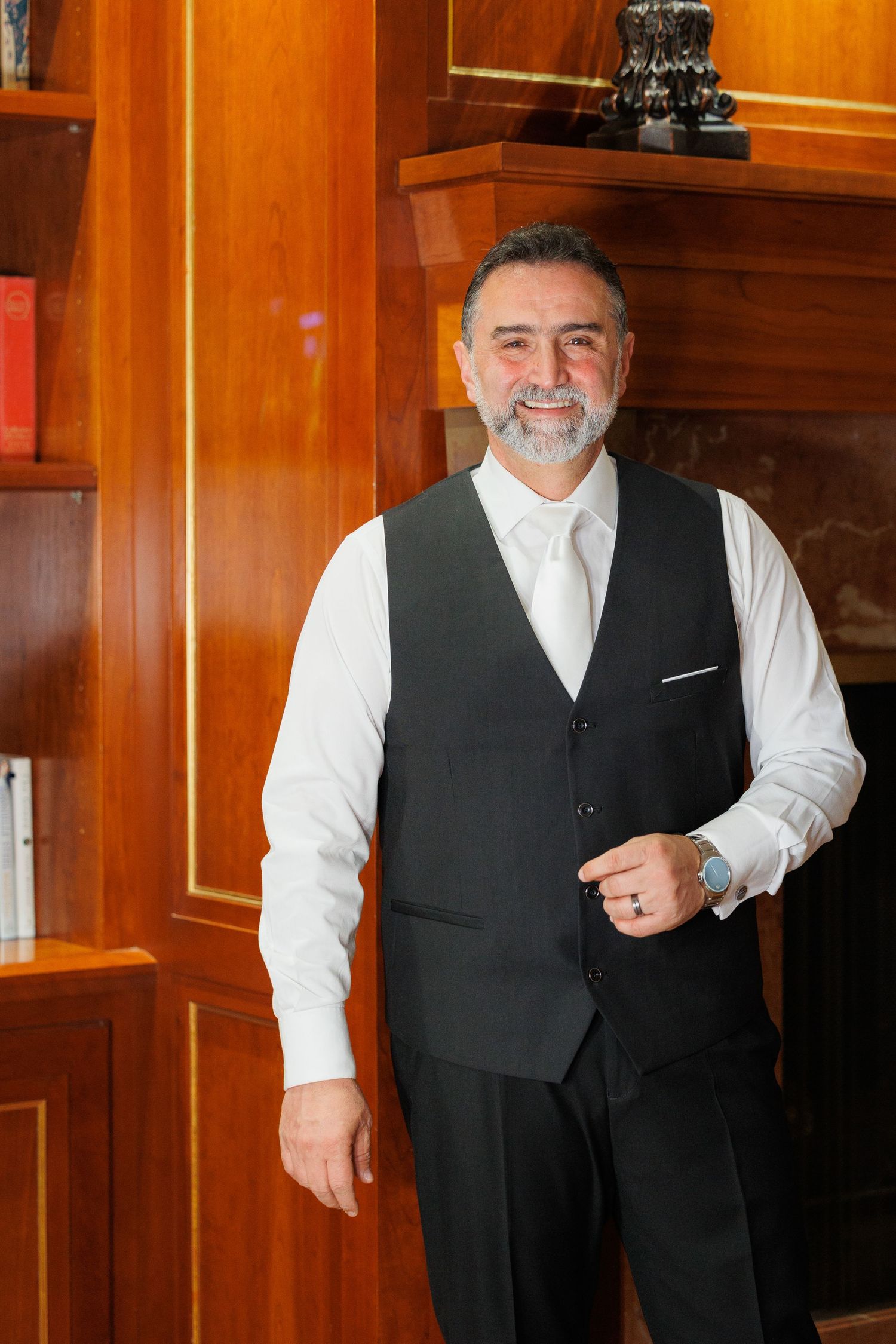 A person in formal attire with a white shirt and black vest stands against a wood-paneled wall in warm lighting.