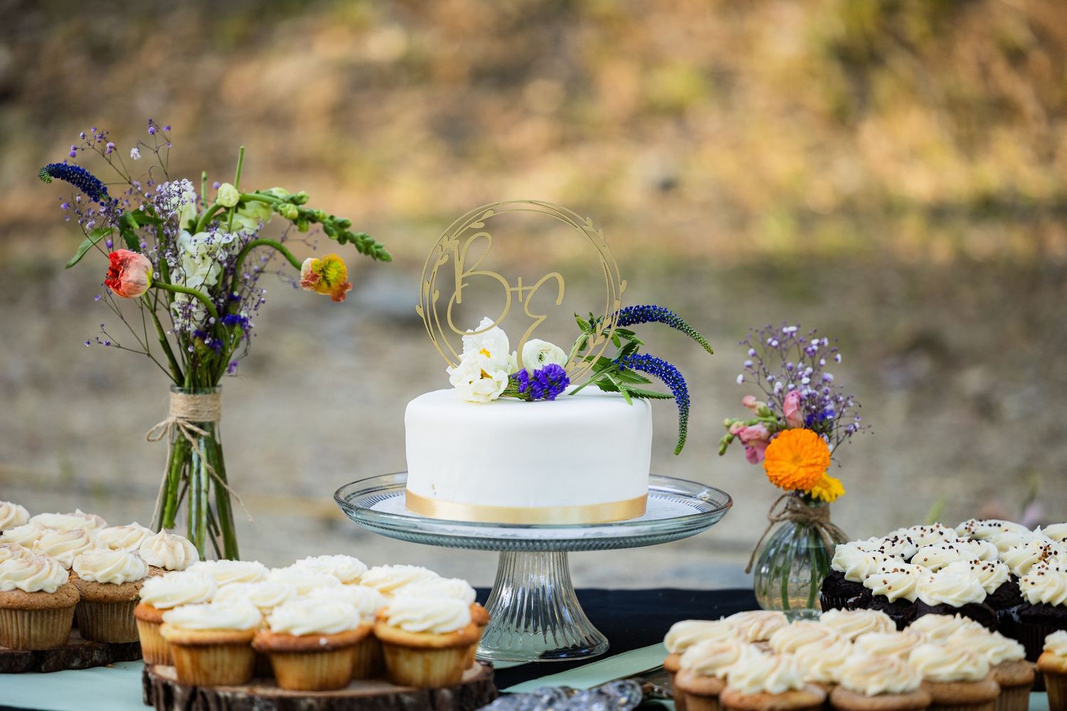 Elegant wedding dessert table display with white cake, cupcakes and floral decorations arranged outdoors.