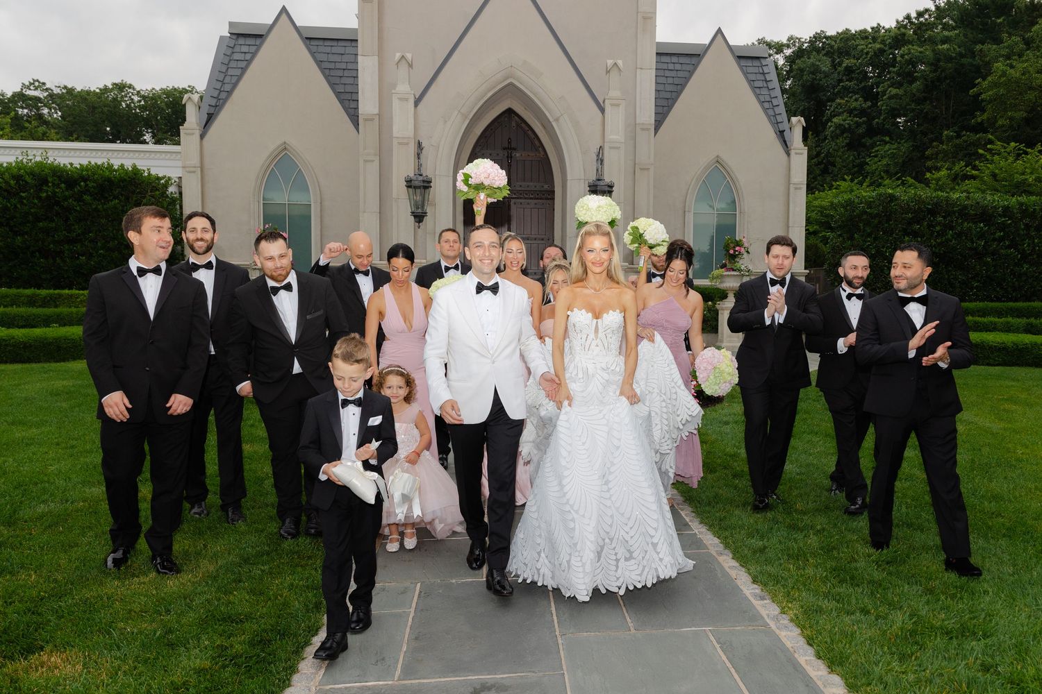 Wedding party members walk and pose together outside a church on a sunny summer day.