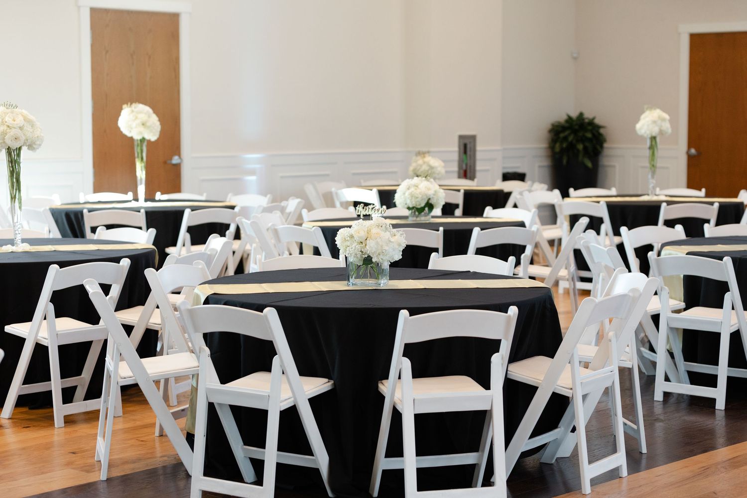 White folding chairs arranged in rows at a banquet venue with black tablecloths and decorative lighting.