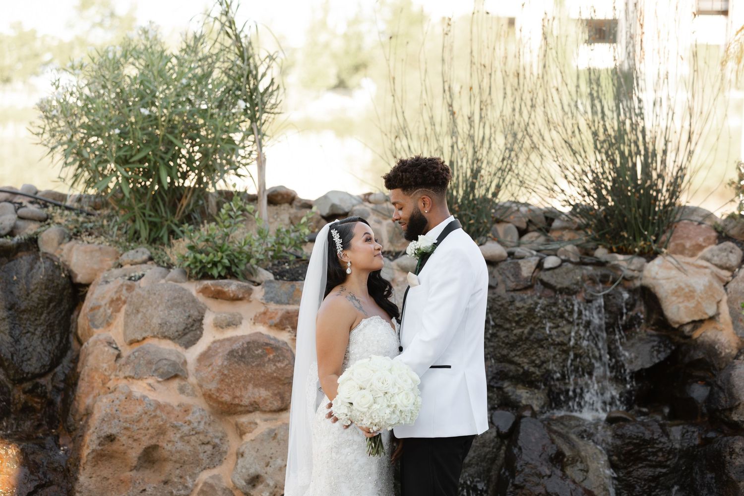 A couple in formal wedding attire poses together against a rustic stone wall backdrop in warm sunlight.