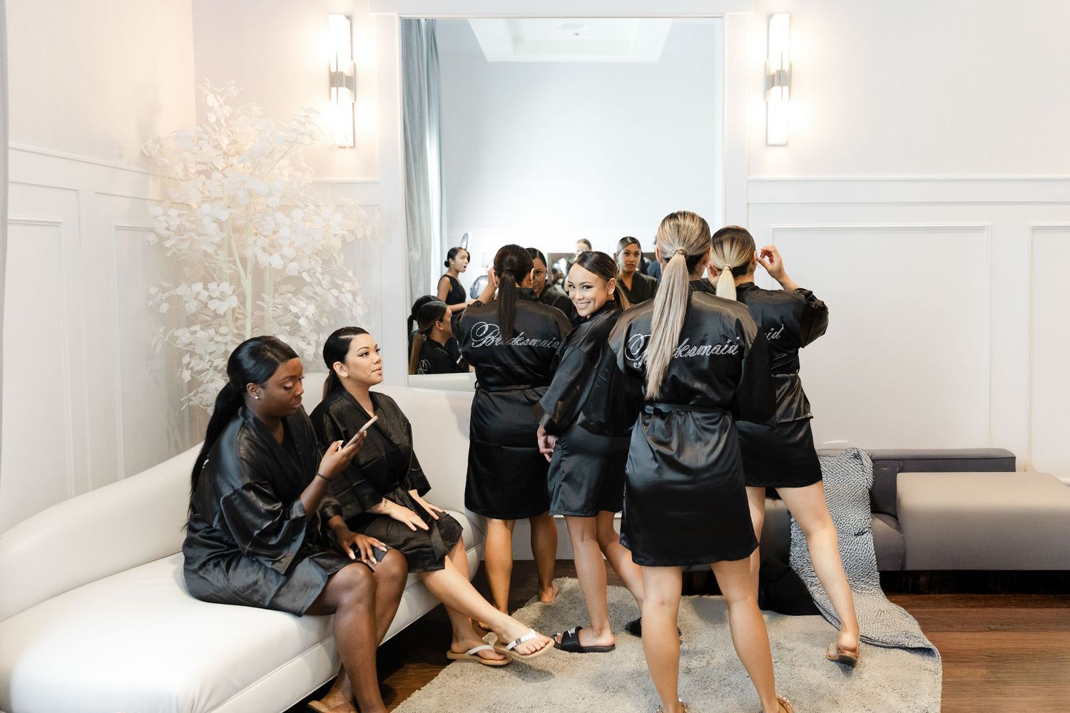A group of dancers stretches and practices together in a bright dance studio with white walls and mirrors.