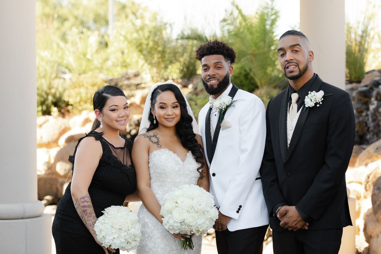 Wedding party dressed in formal black and white attire laughs together while posing outdoors on sunny day.