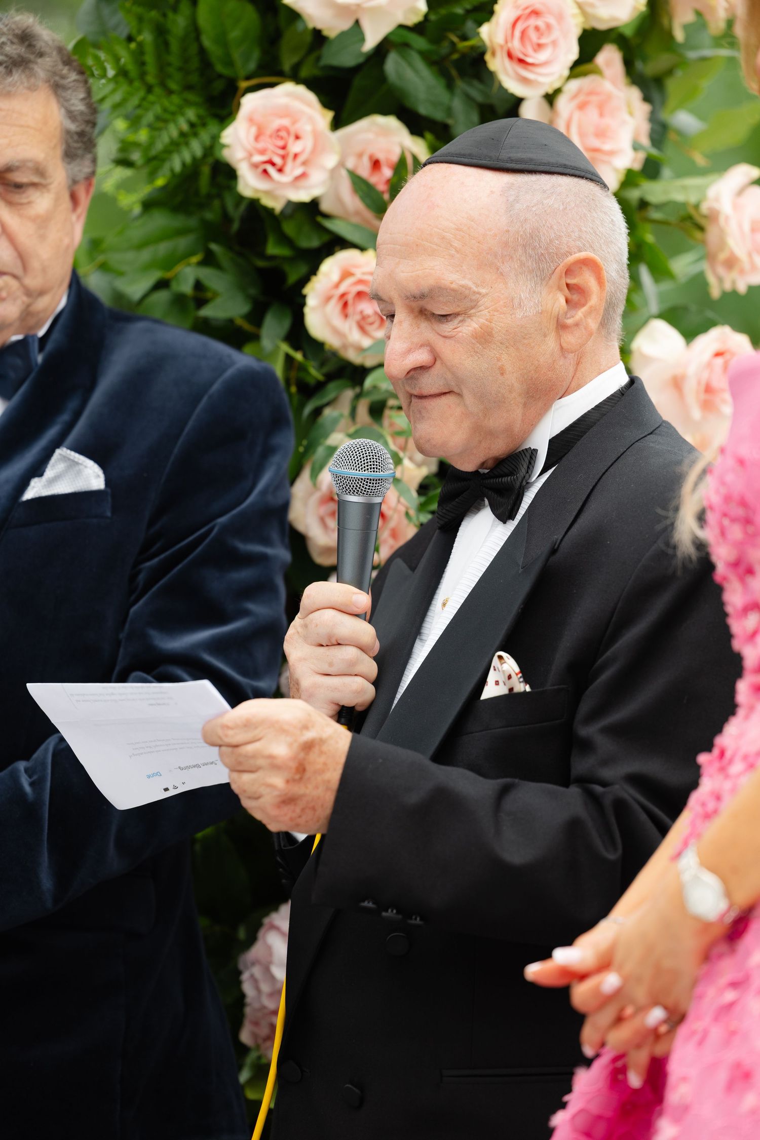 A speaker in a black tuxedo holds a microphone while reading from notes at a formal event with pink floral decor.