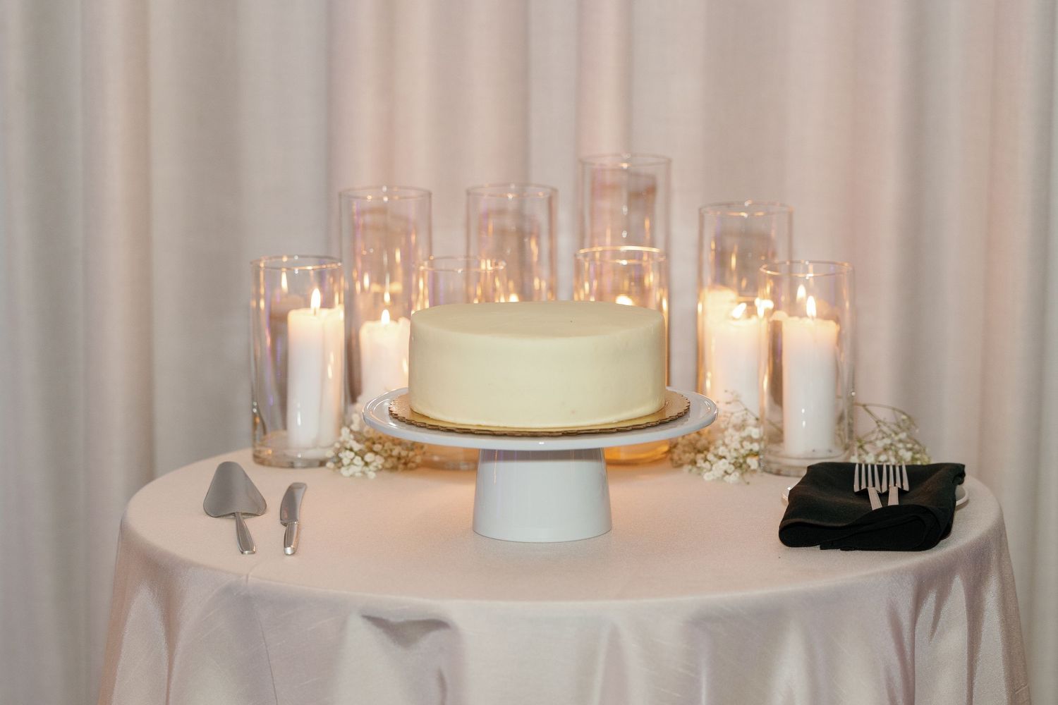 Simple white wedding cake on a pedestal surrounded by glowing glass candles on an elegant white tablecloth.