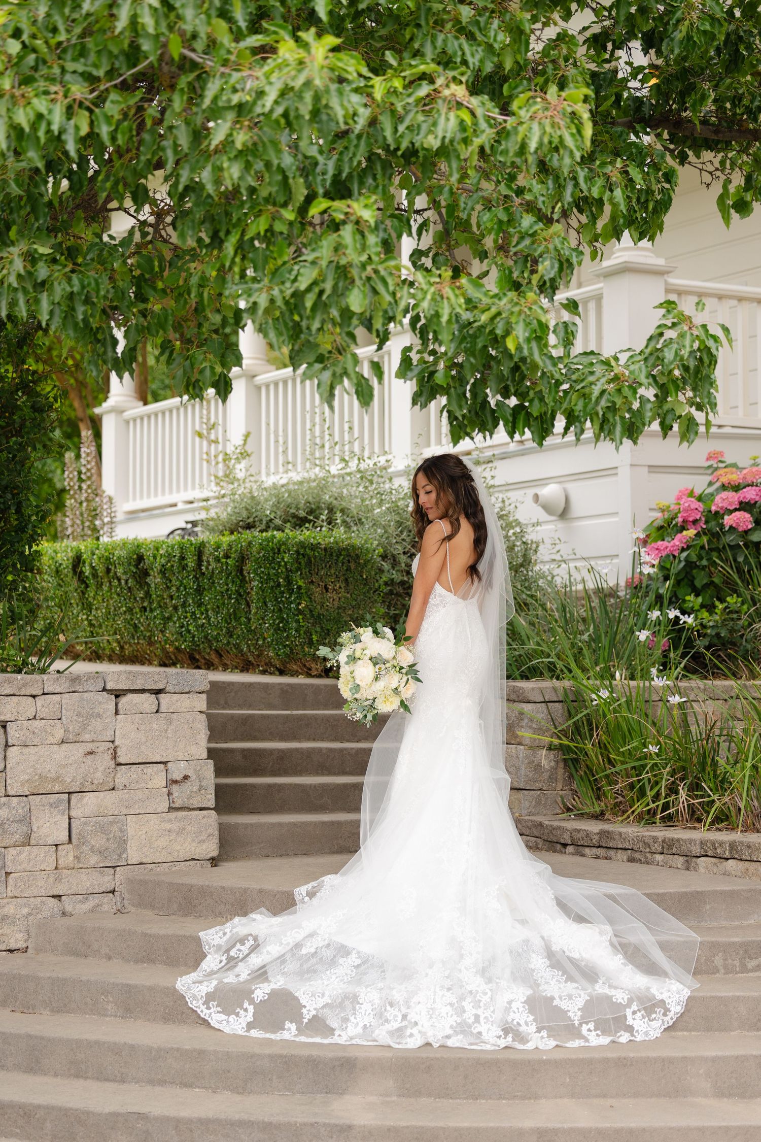 A bride in a flowing white wedding gown with lace train sits gracefully on stone steps under green foliage.