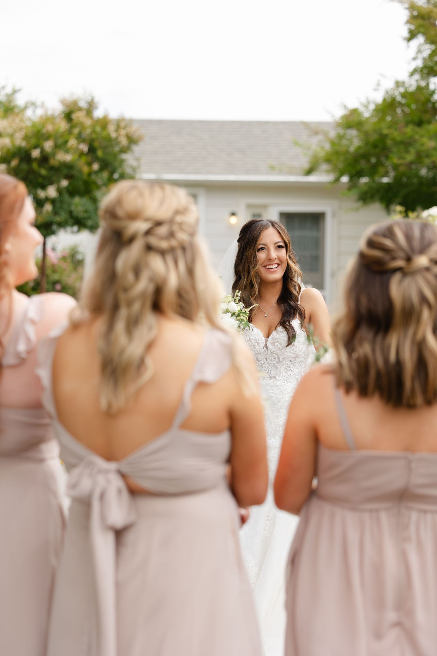 Bridesmaids in blush pink dresses stand in a row outdoors at a wedding celebration.