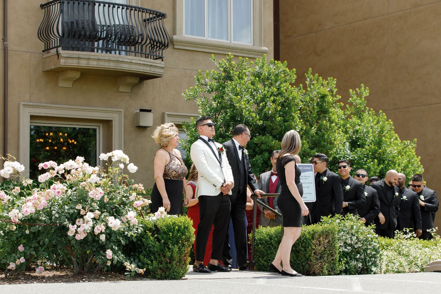 People gather outside a building with wrought iron balconies and blooming rose bushes along a walkway.