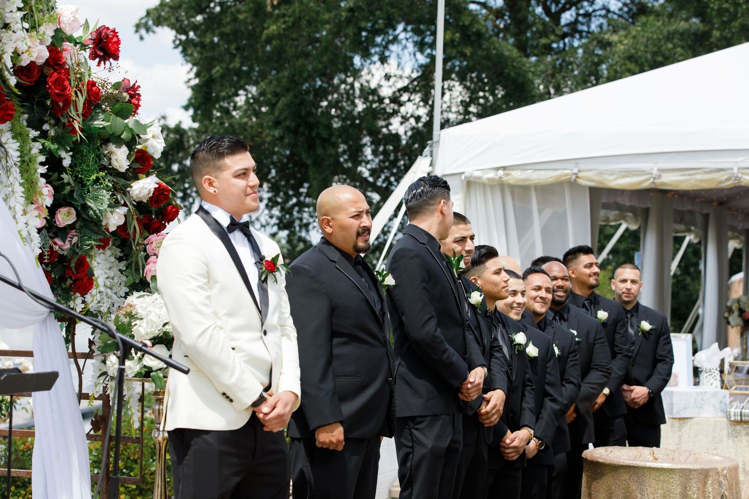 A wedding party poses together under white tents and floral decorations at an outdoor celebration.