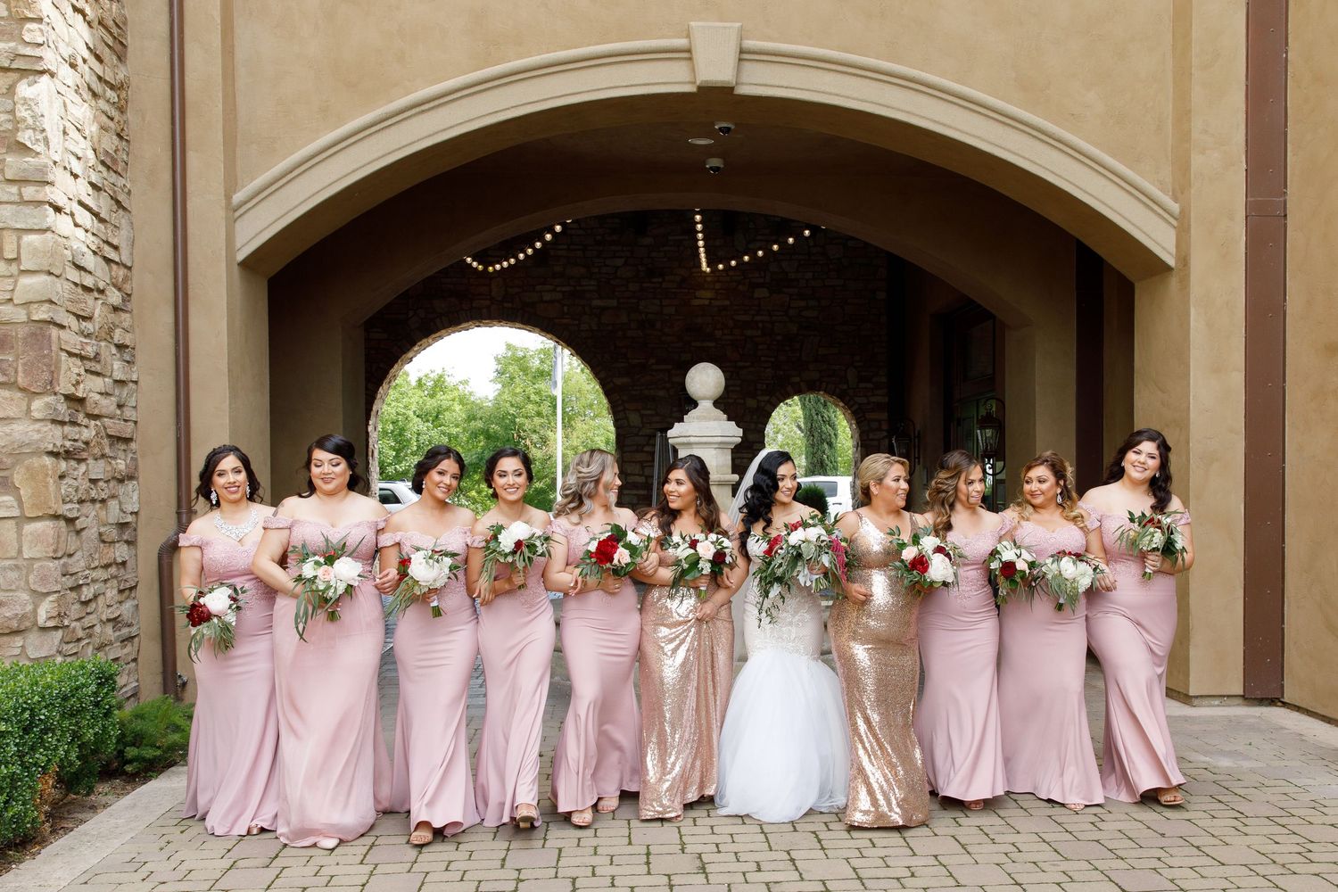 Wedding party in pink bridesmaid dresses and white bridal gown pose under stone archway with floral decorations.