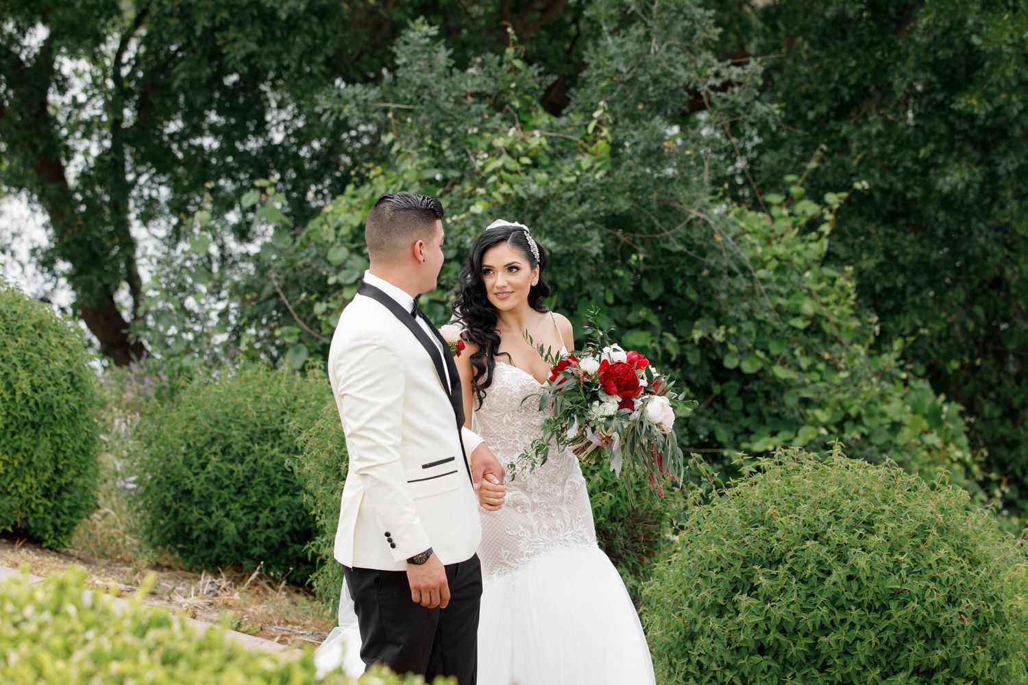 Newlywed couple stands together outdoors with red bouquet against lush green garden backdrop on their wedding day.