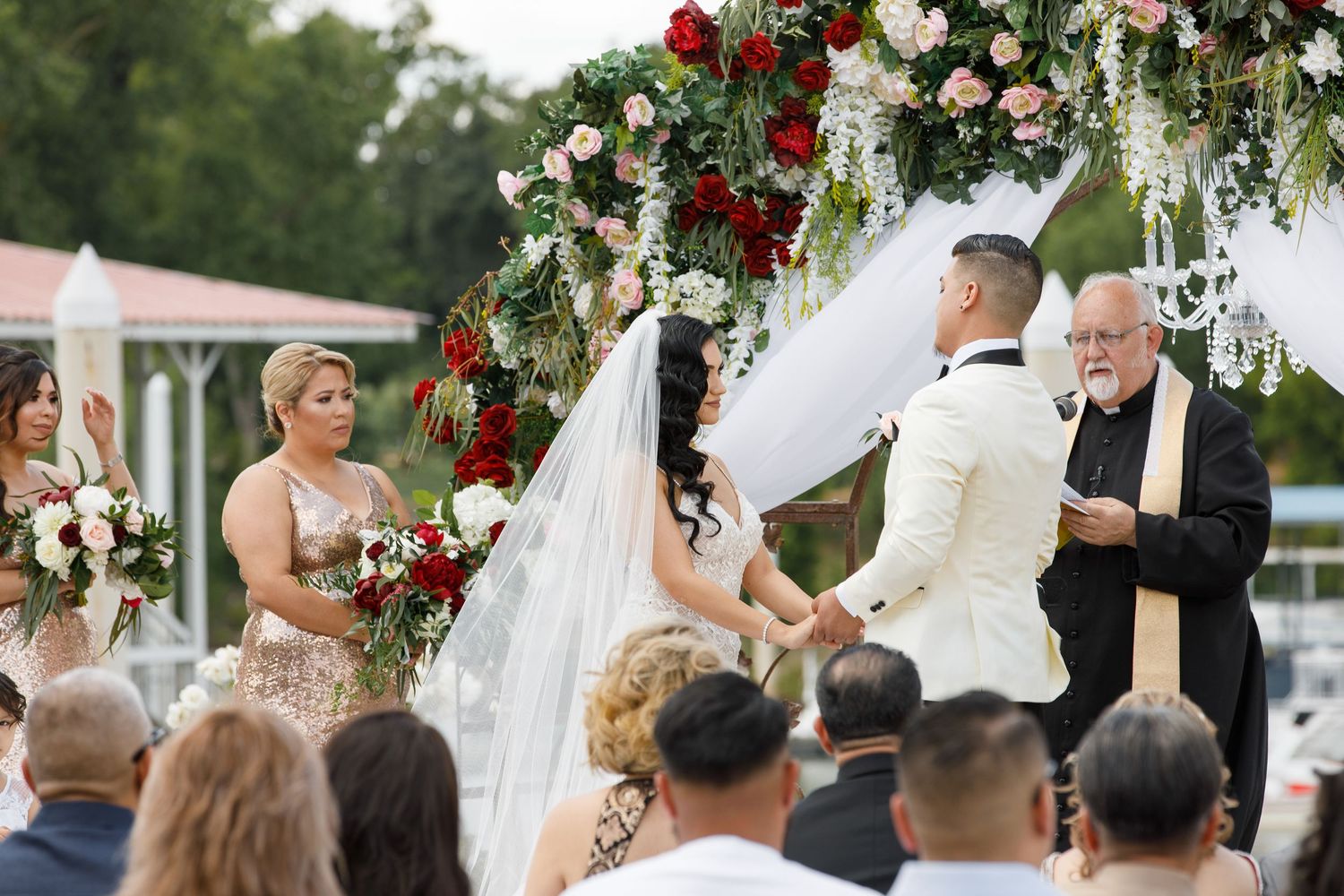 An outdoor wedding ceremony under a floral arch decorated with red and white roses and greenery.