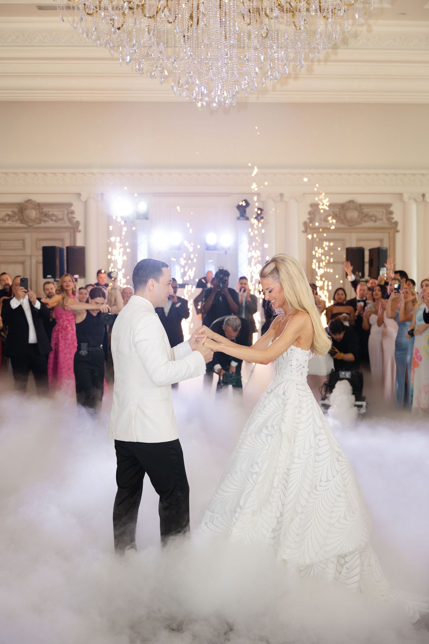 A couple shares their first dance at an elegant wedding reception with fog effects and a crystal chandelier overhead.
