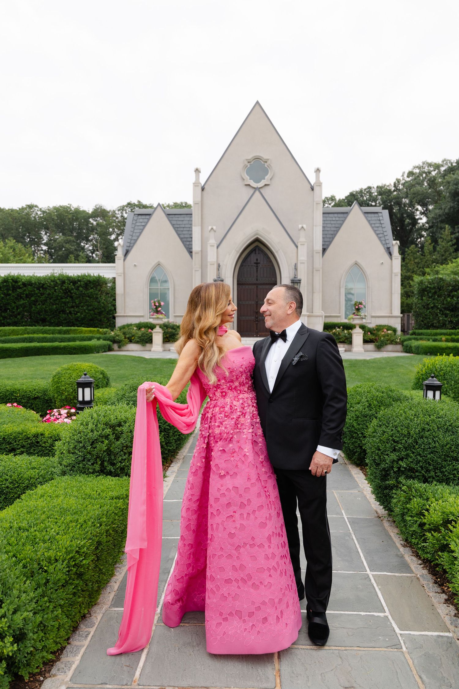 A couple walks along a pathway leading to a white gothic-style chapel surrounded by manicured hedges.