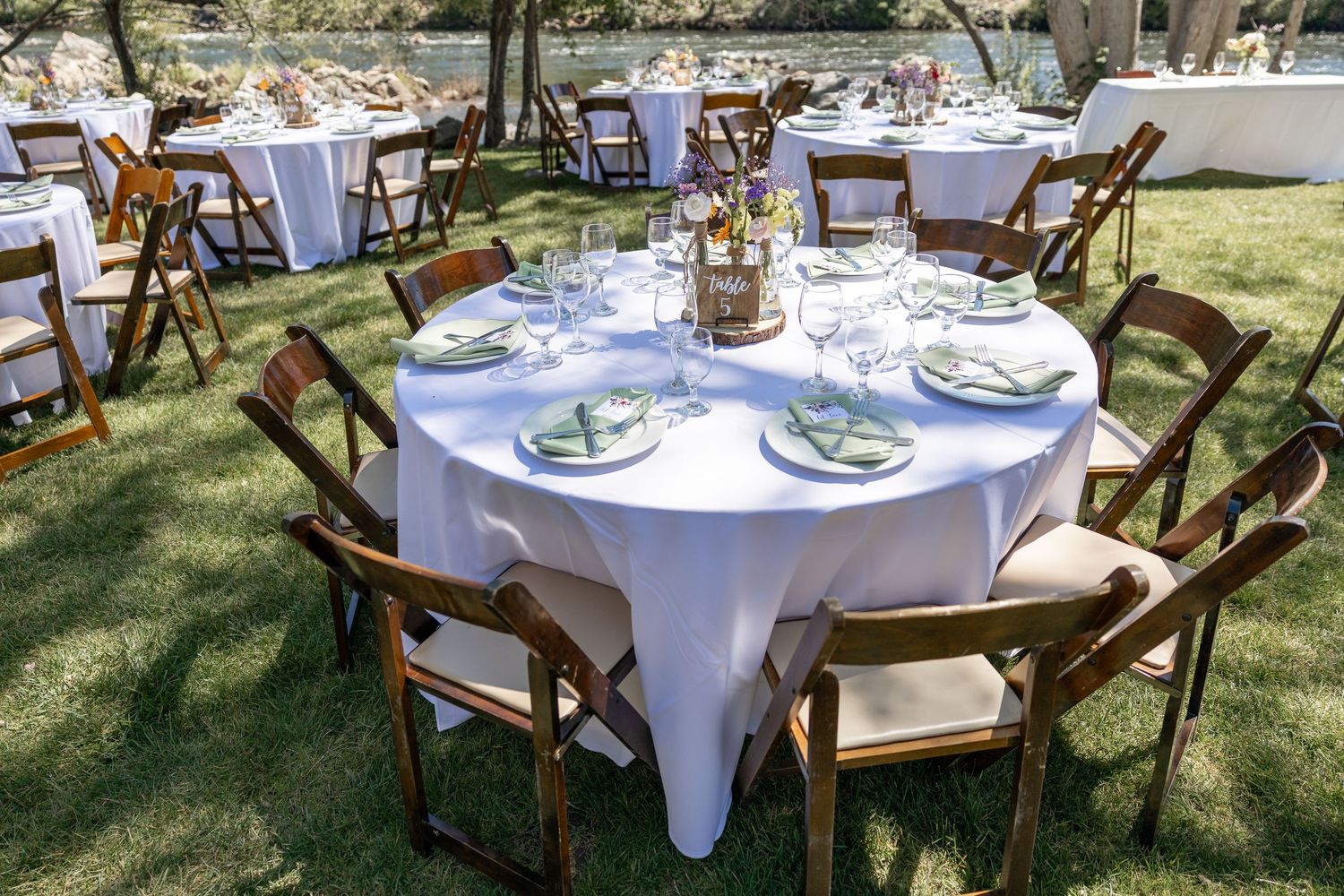 Elegant outdoor wedding reception setup with round tables, white linens, and rustic wooden cross-back chairs in a garden setting.