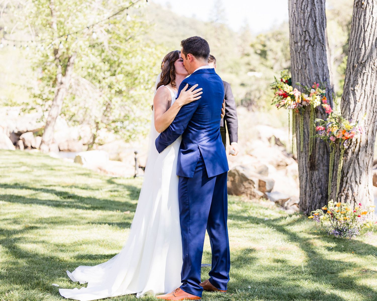 A romantic wedding couple shares tender moments in a sun-dappled outdoor garden setting with tall trees.