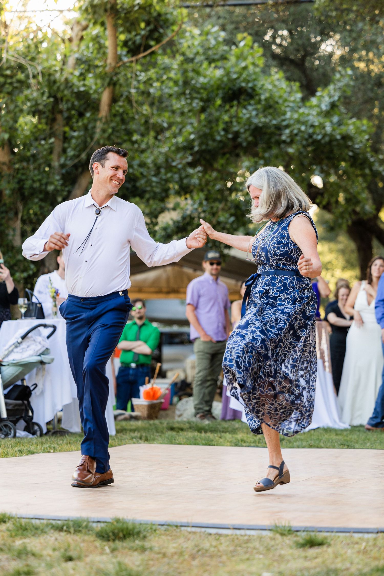 Two people dancing joyfully together outdoors at a summer gathering, with one wearing a blue patterned dress.