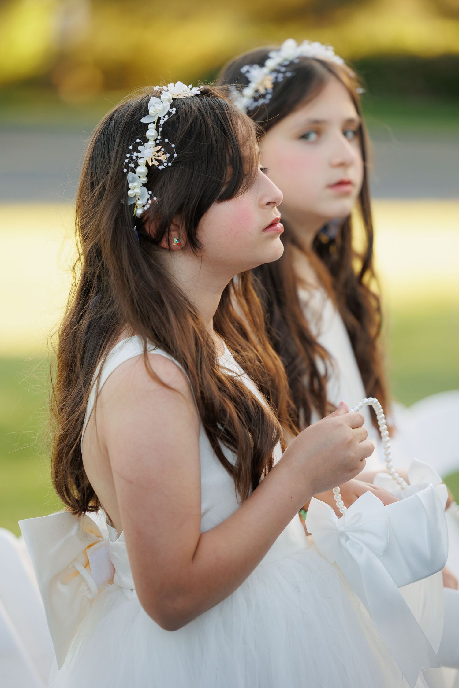 Two flower girls in white dresses with delicate floral headpieces and long wavy hair at an outdoor wedding ceremony.
