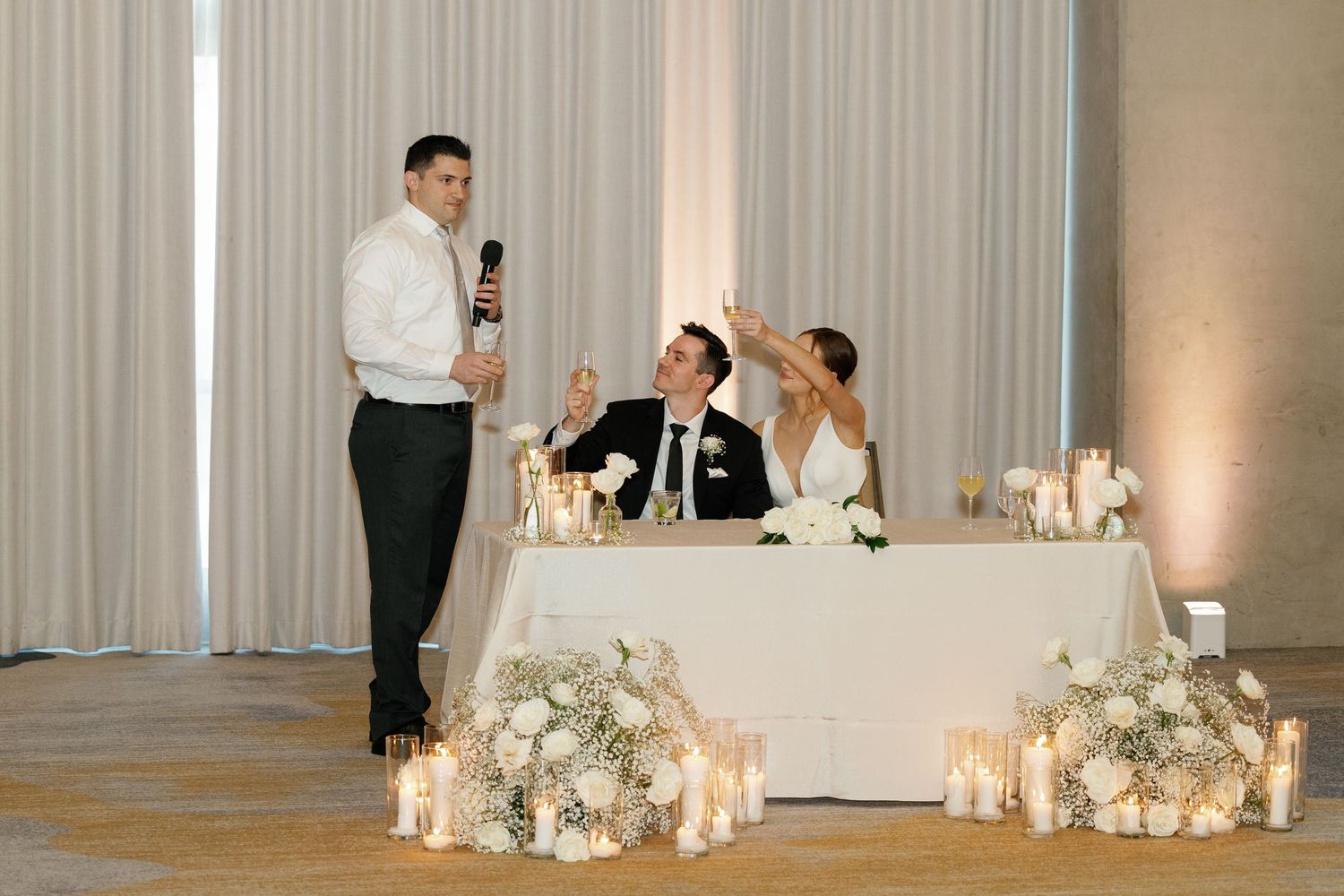 Elegant wedding reception setup with white floral centerpieces, glowing candles and draped curtains creating a romantic atmosphere.