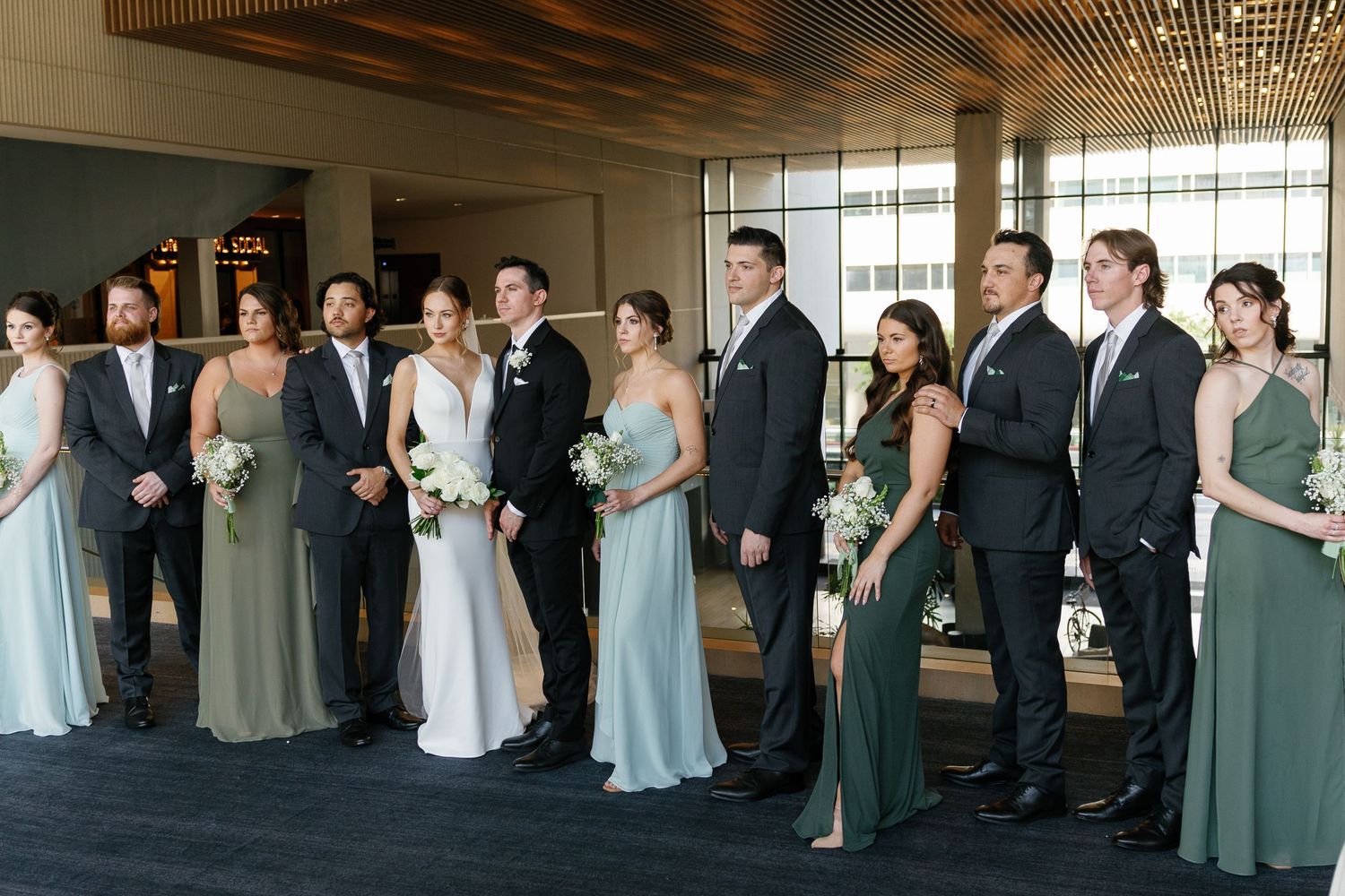 Wedding party members lined up in formal attire with bridesmaids in sage green dresses and groomsmen in dark suits.