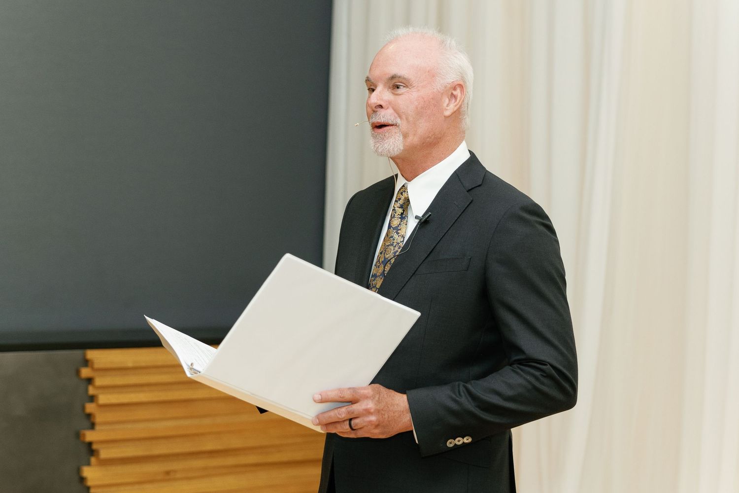A speaker in a dark suit holds papers while presenting at a podium.