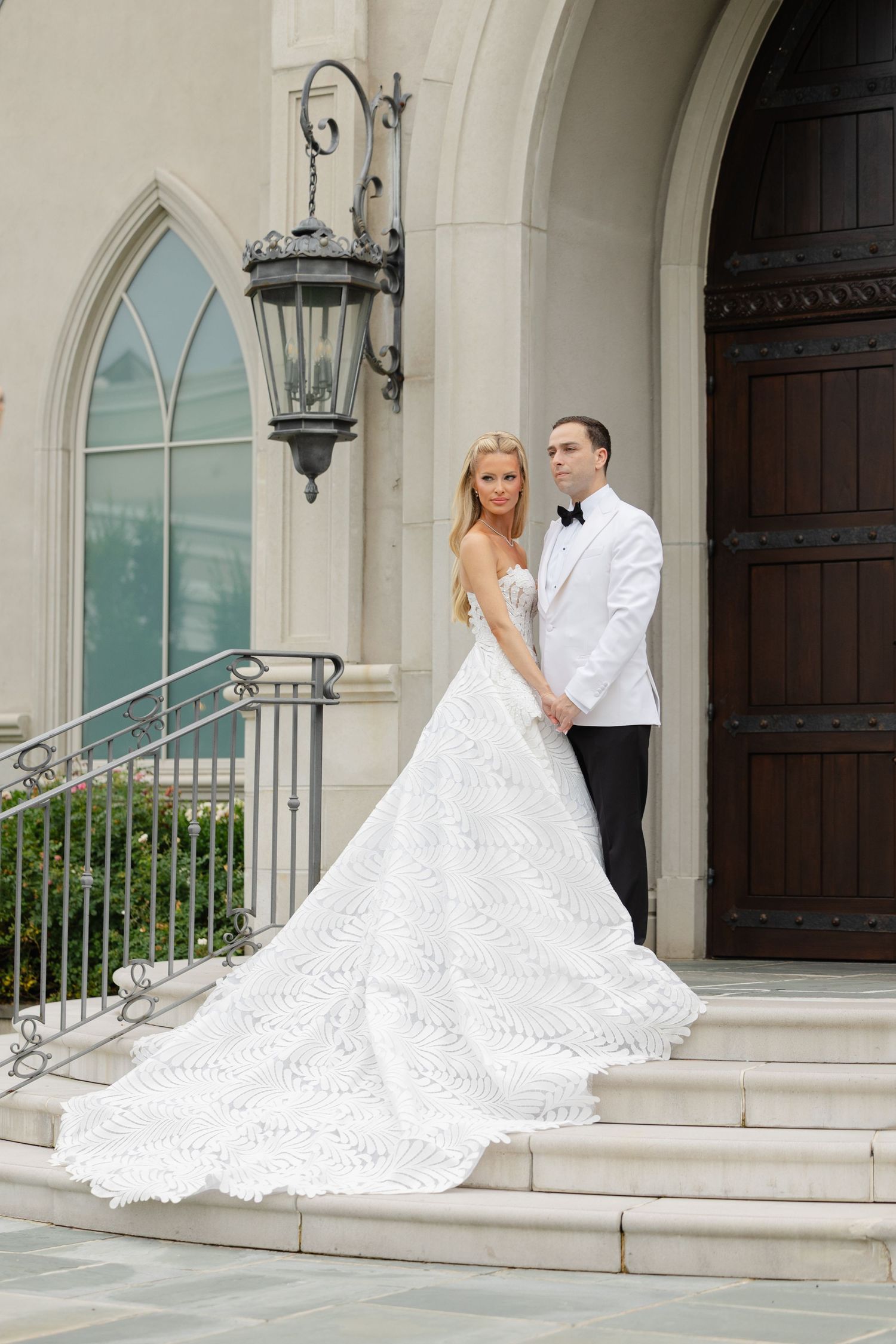 A couple poses on church steps with ornate architecture, featuring a flowing white wedding gown and classic tuxedo.