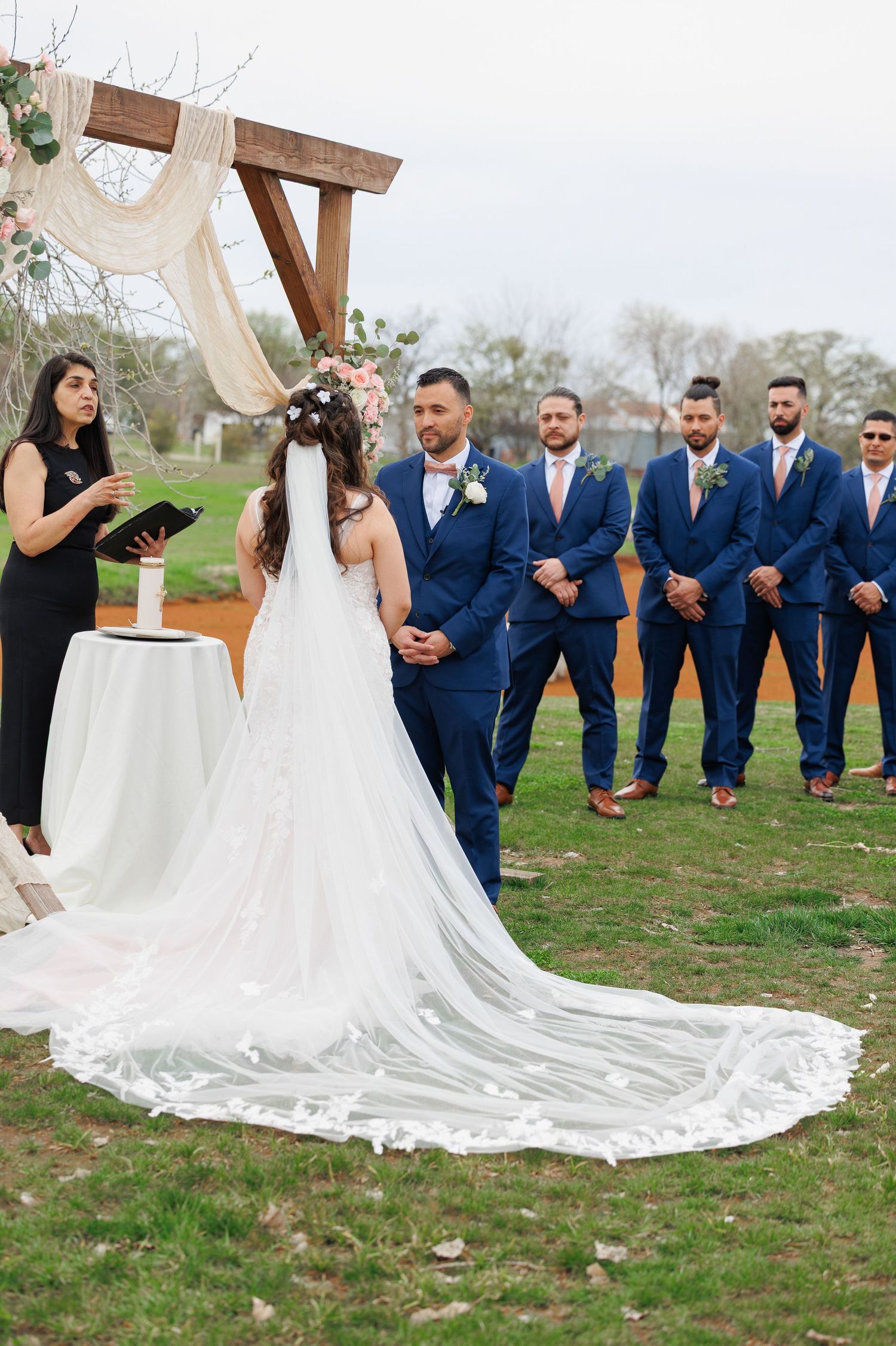 Wedding party in navy blue suits kneels behind bride's flowing white cathedral length train in outdoor garden setting.
