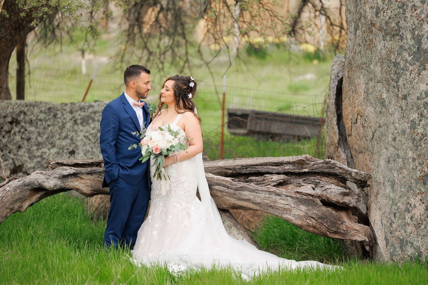 Newlywed couple embraces near rustic fallen tree in green meadow during outdoor wedding photoshoot.