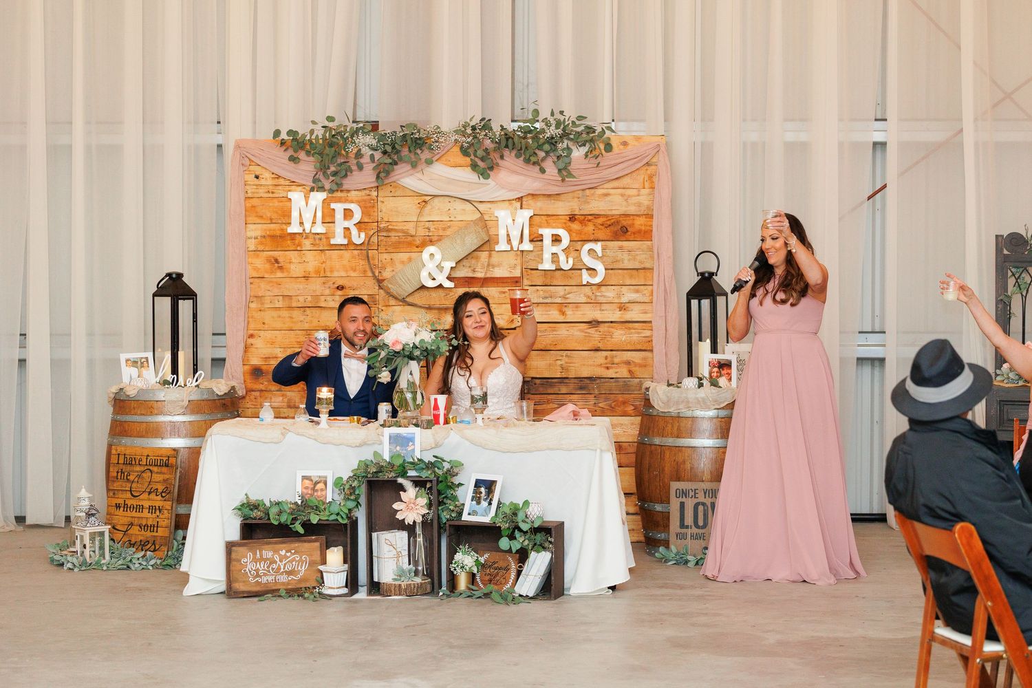 Wedding party sits at a rustic wooden head table decorated with greenery and Mr & Mrs signage during reception.