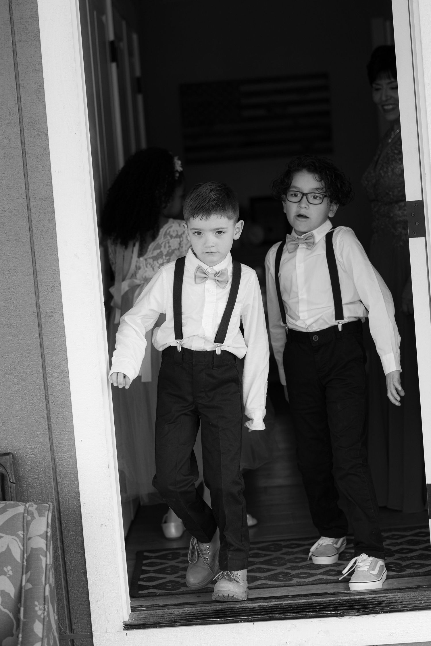 Two children in formal black and white attire with suspenders and bow ties stand in a doorway.