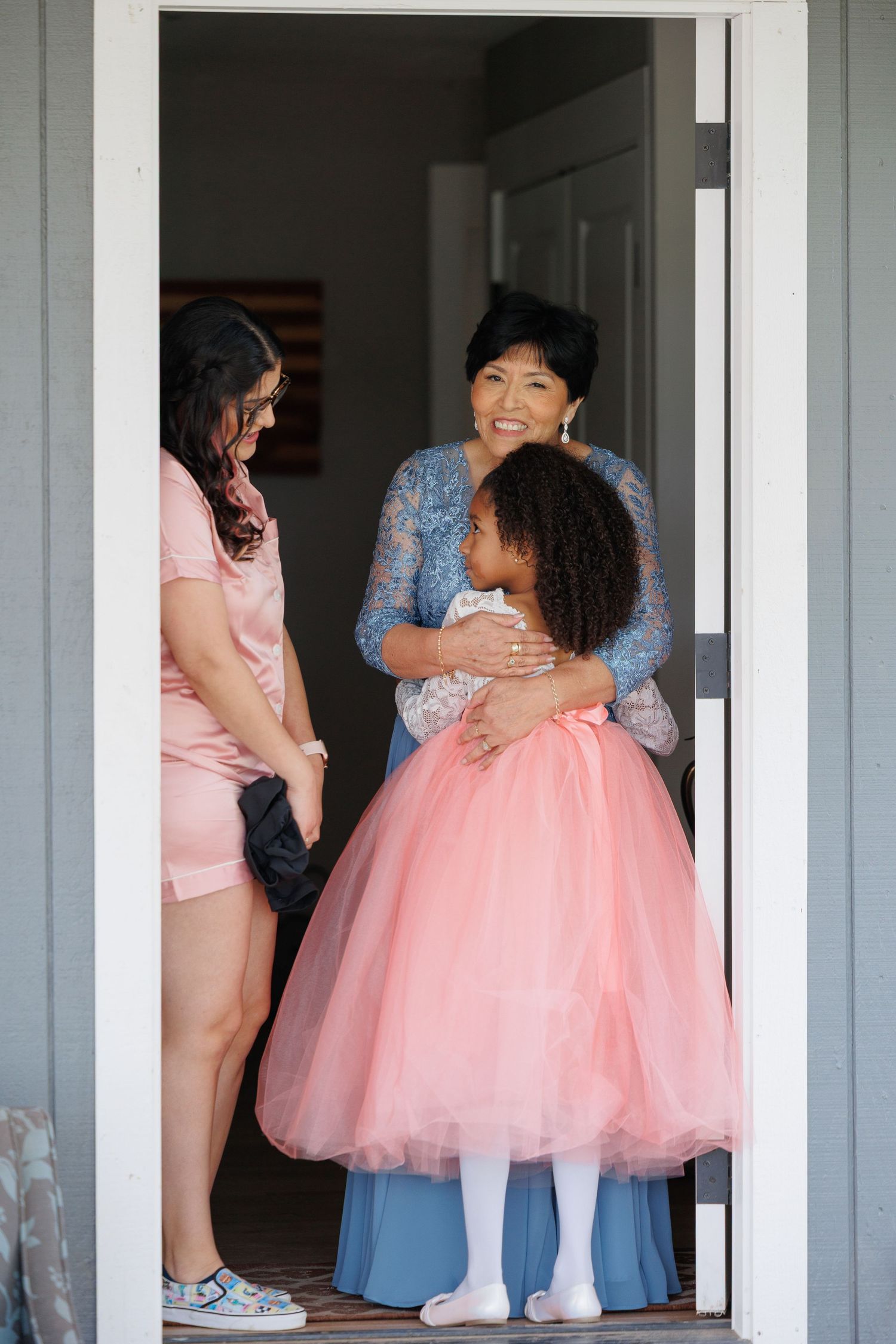 A person in a blue dress helps adjust a flowing pink tulle gown in a white hallway.