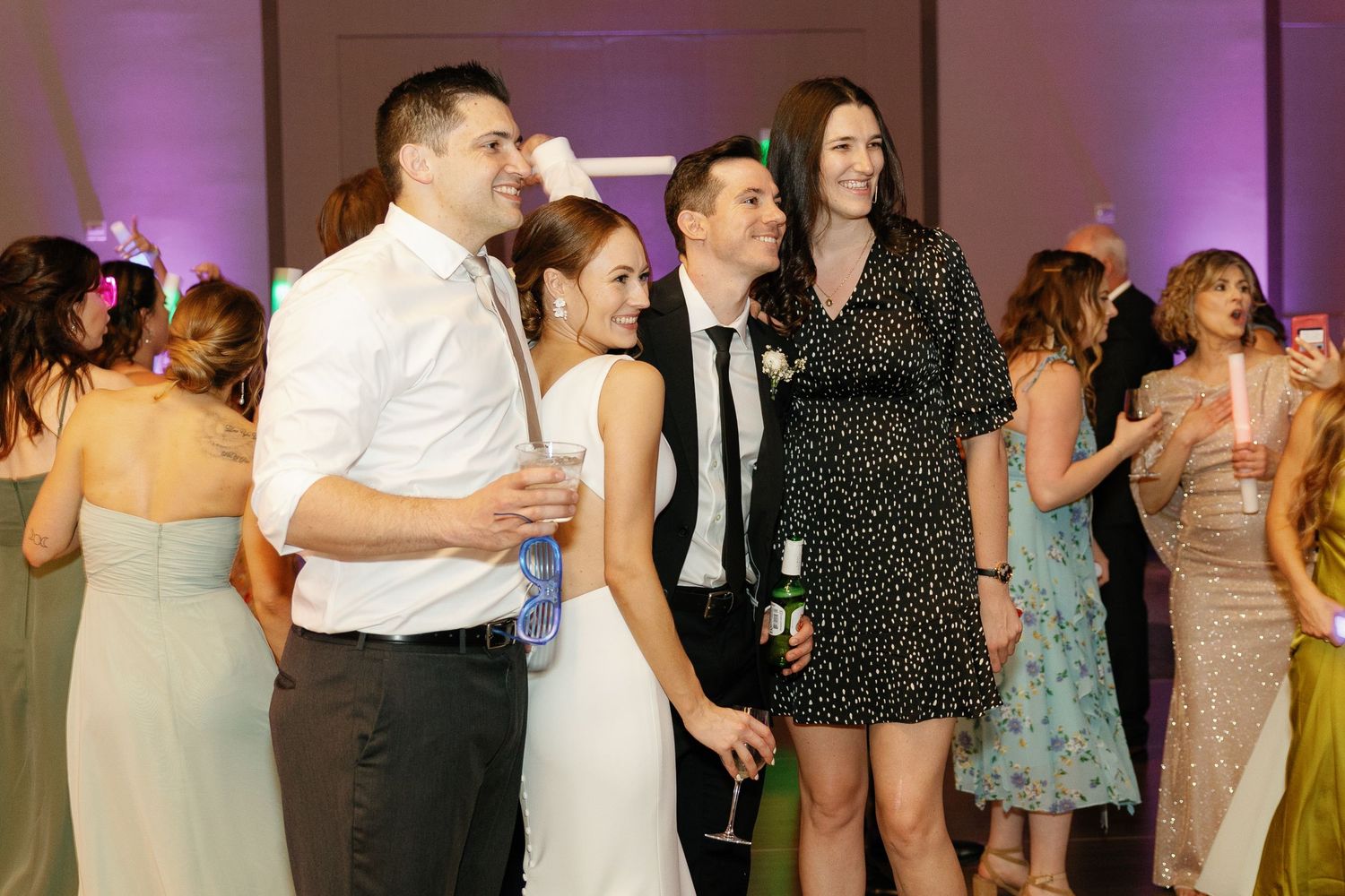 Wedding guests dance together on a crowded dance floor under purple lighting at an evening reception celebration.