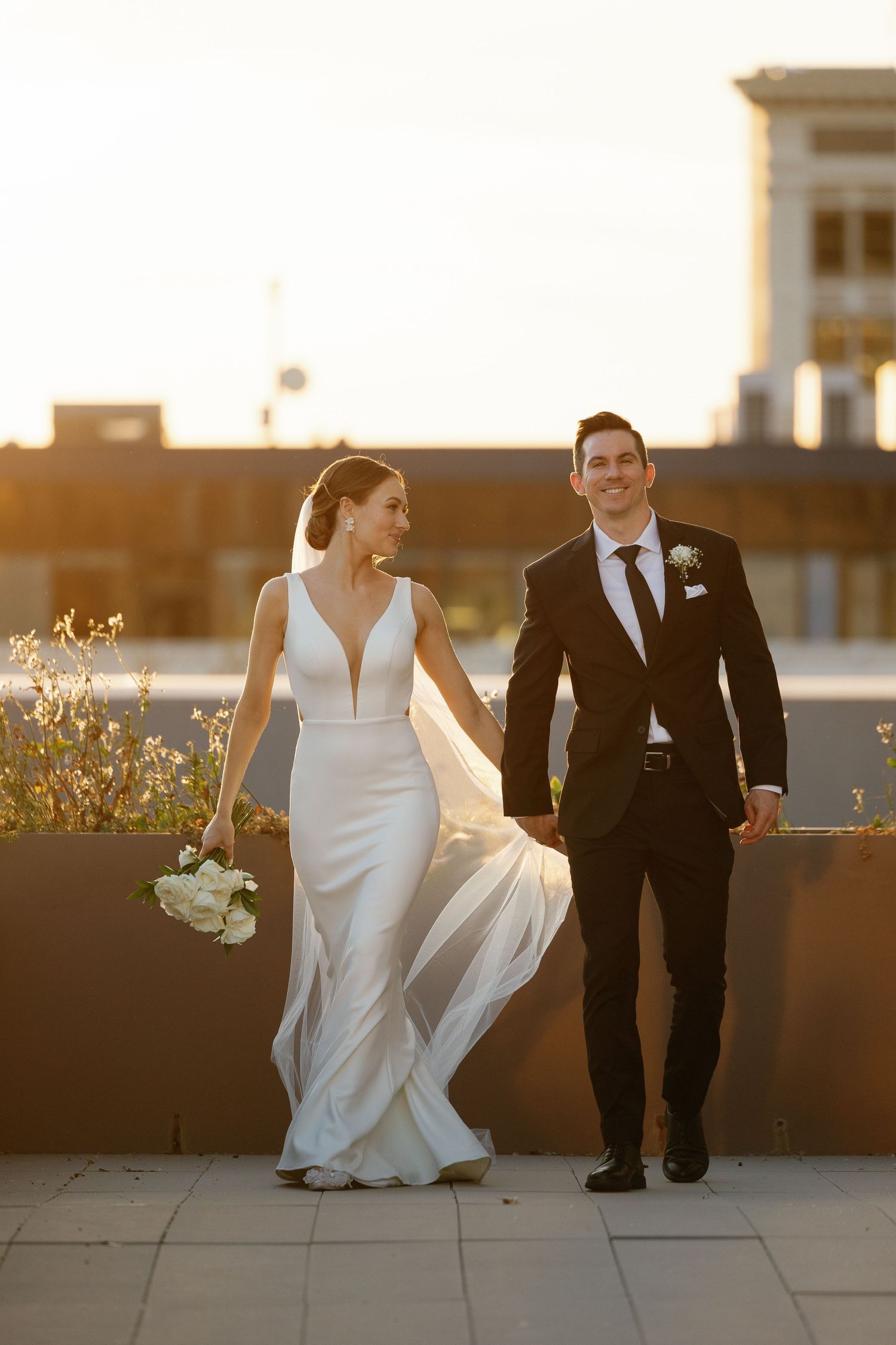 A bride in a sleek white gown and groom in black suit walk together during golden hour sunset on a rooftop.