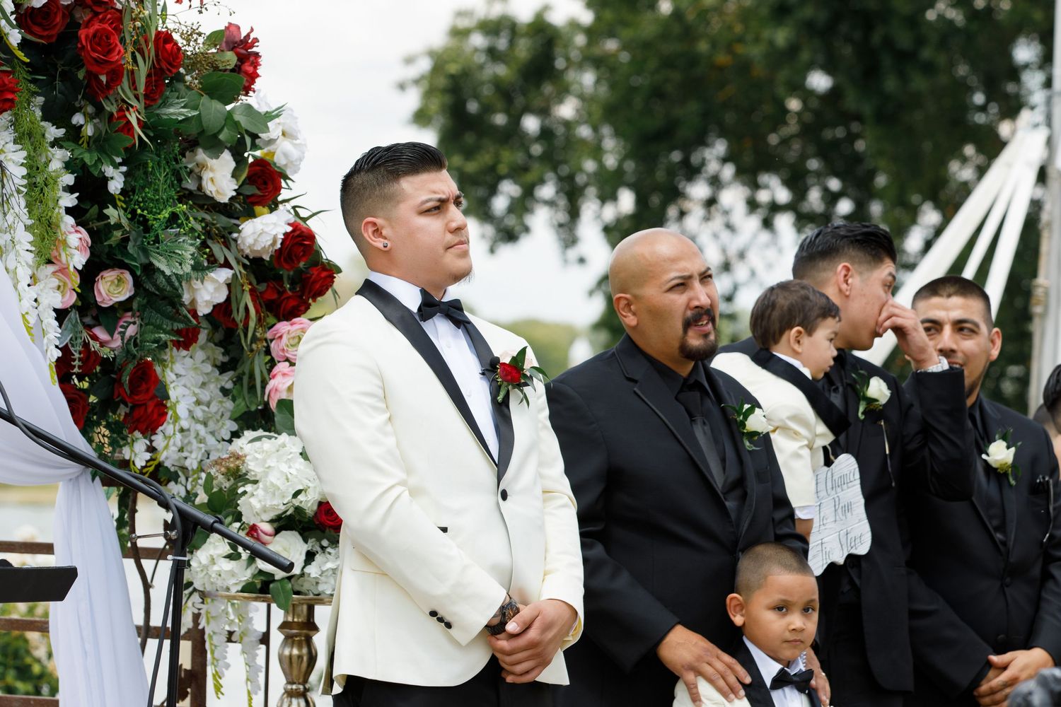 Wedding party gathers outdoors under floral arch decorations with red roses and white flowers during ceremony.