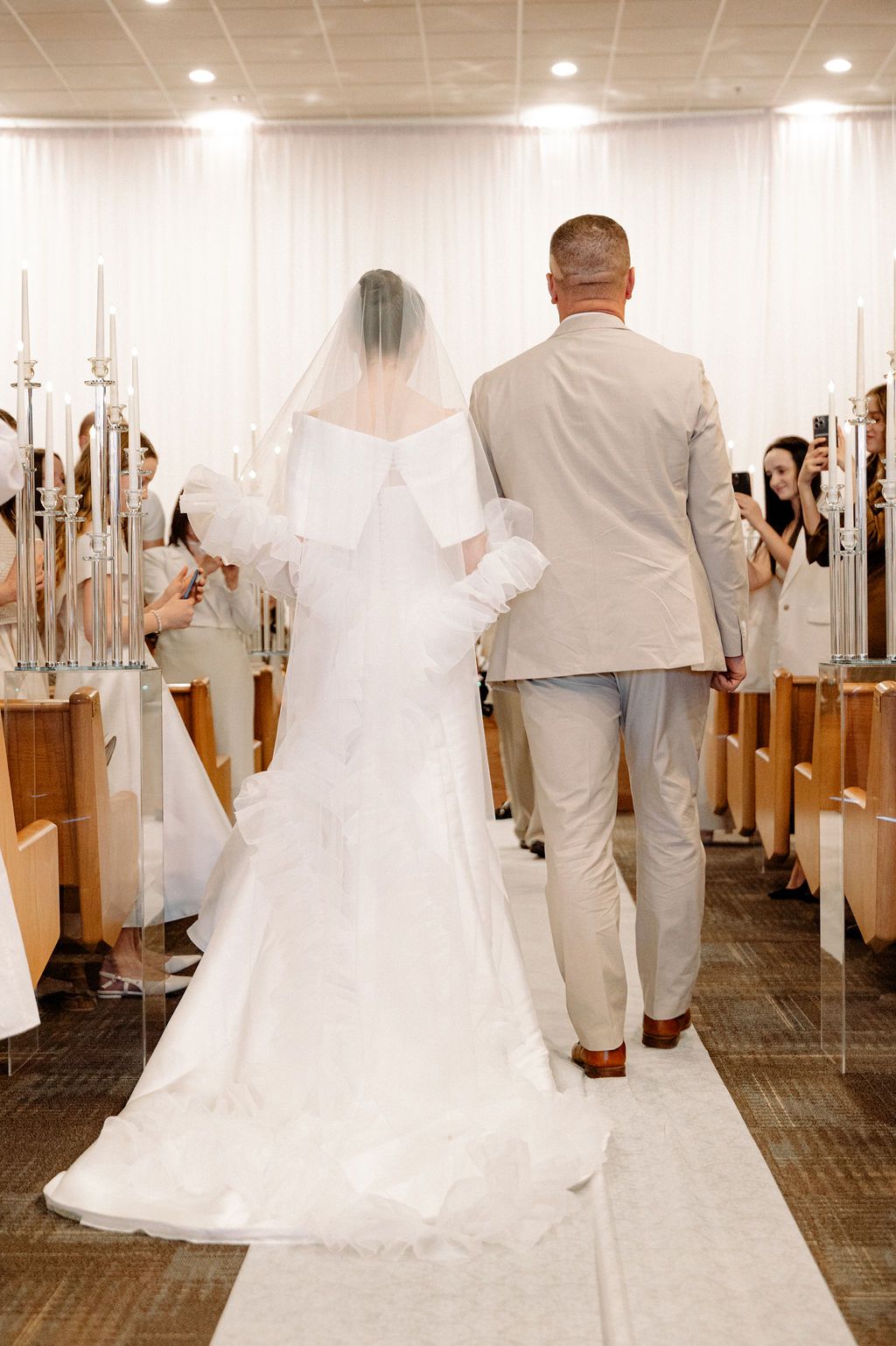 A wedding ceremony processional showing a couple walking down the aisle with white decor and candles.