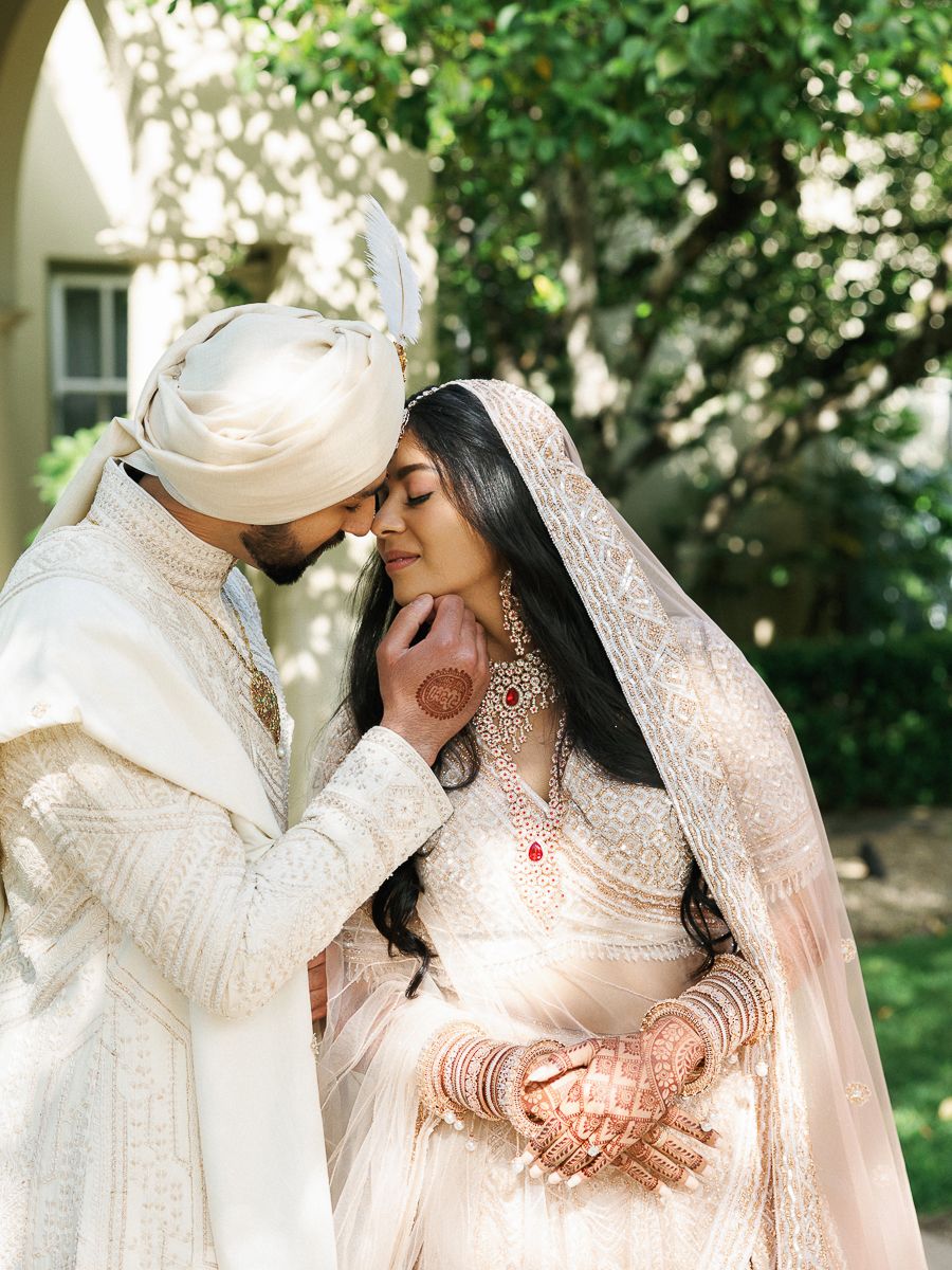 A romantic wedding couple in traditional white and cream South Asian attire share intimate moments in a garden setting.