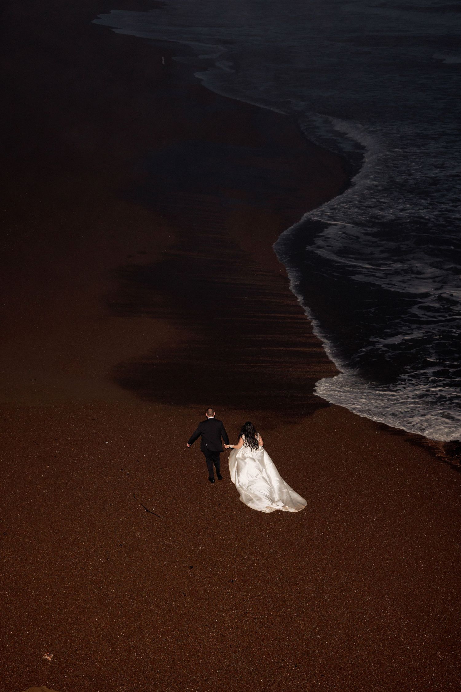 A couple walks along a dark sandy beach at sunset with white waves lapping at the shoreline.