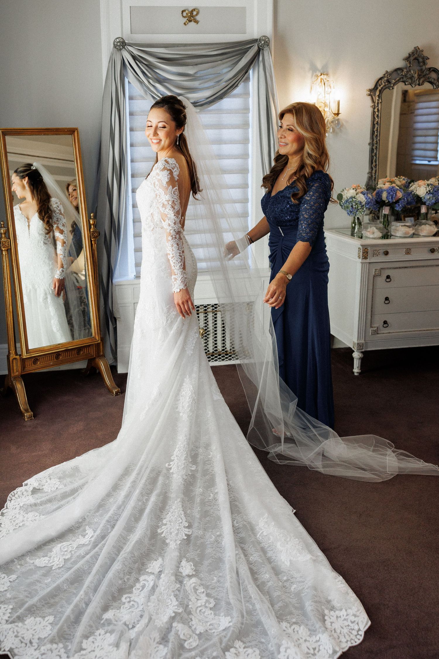 A stunning lace wedding gown with a dramatic train being fitted in an elegant dressing room with mirrors.