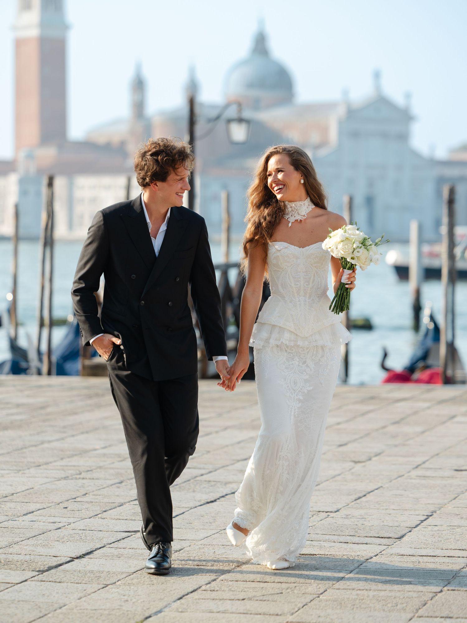 A couple in wedding attire walks along Venice's historic waterfront with the iconic San Giorgio Maggiore church in background.