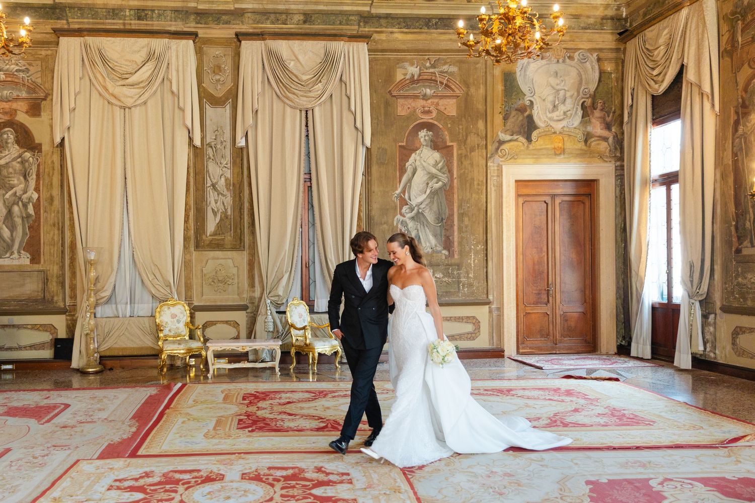 A couple dances in an ornate ballroom with gold chandeliers, marble columns and elegant drapes.