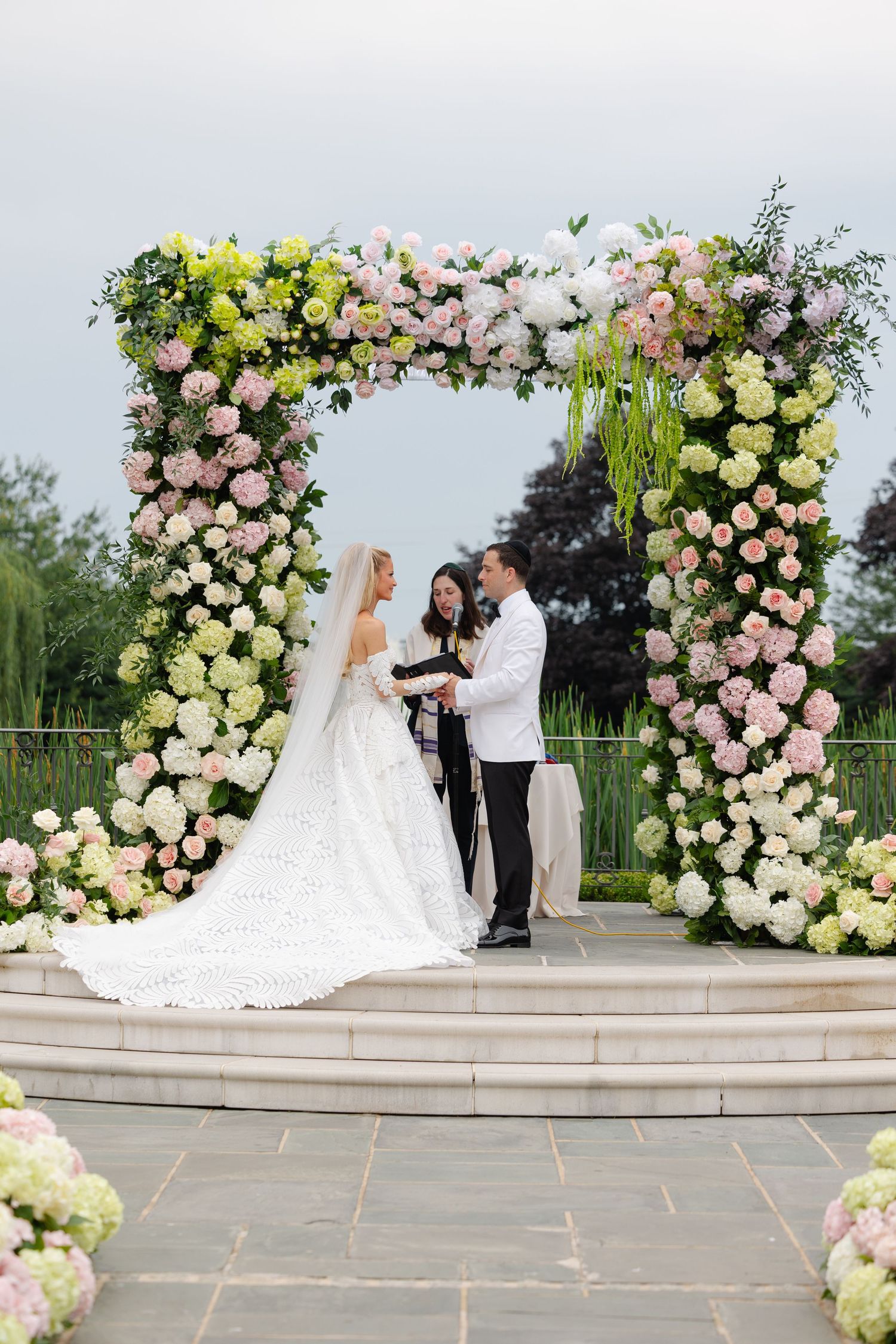 Wedding ceremony takes place under an elegant floral arch with pink and white roses on stone steps outdoors.