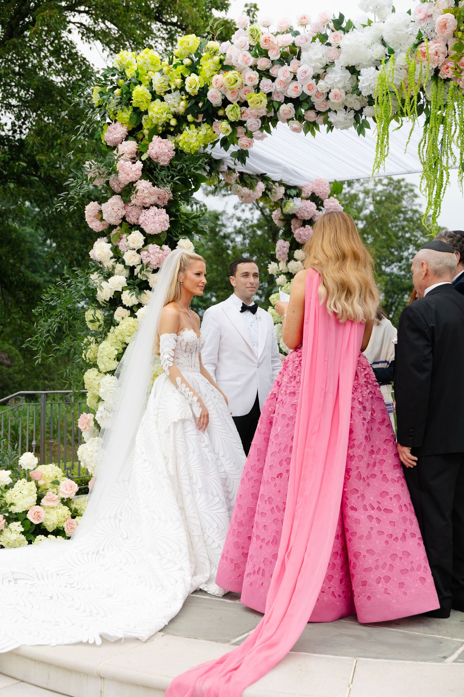 Wedding party gathers under a floral arch with pink and white decorations during an elegant outdoor ceremony.