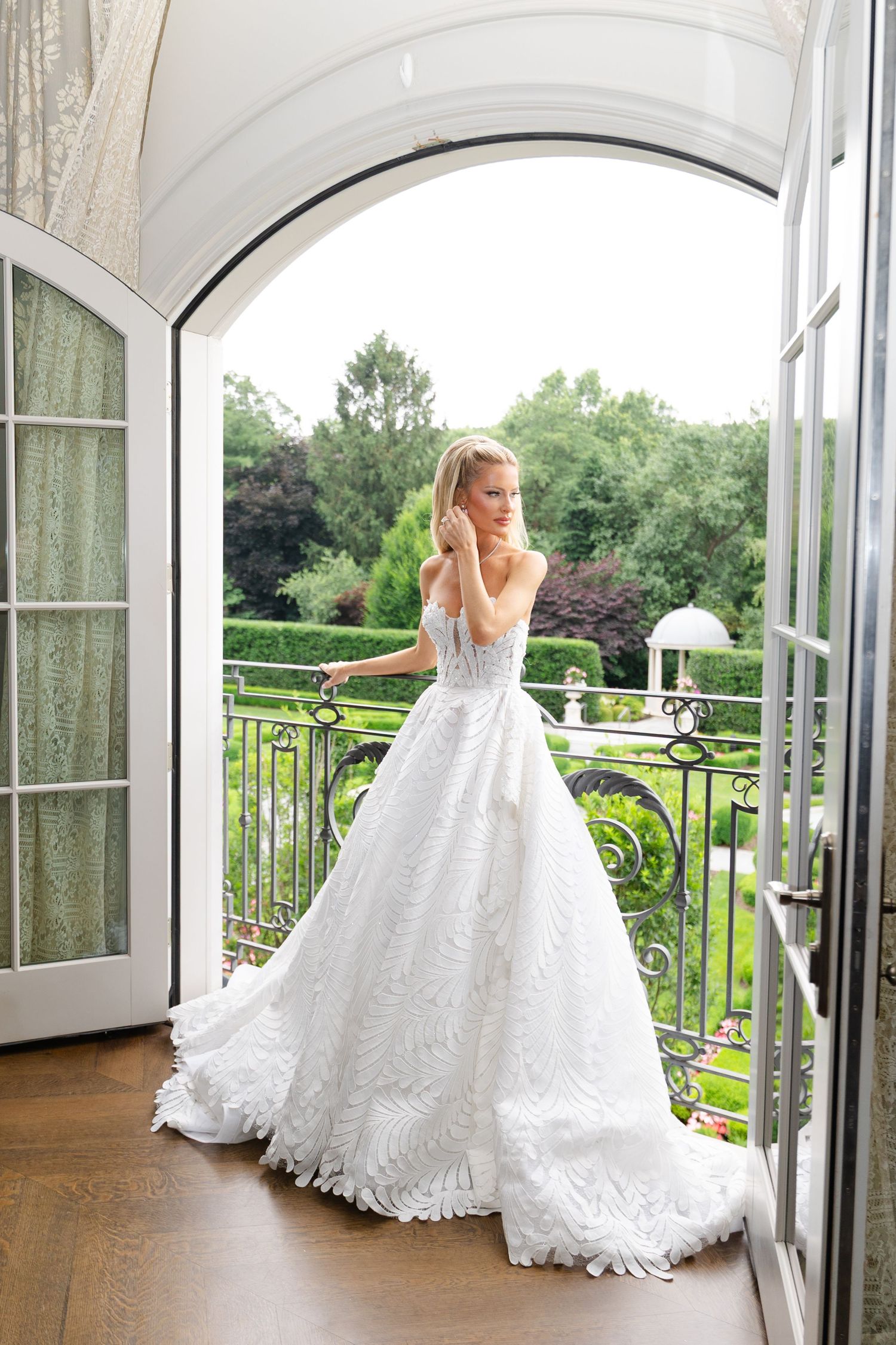 A bride in a white lace wedding dress stands gracefully by French doors overlooking a scenic garden balcony view.