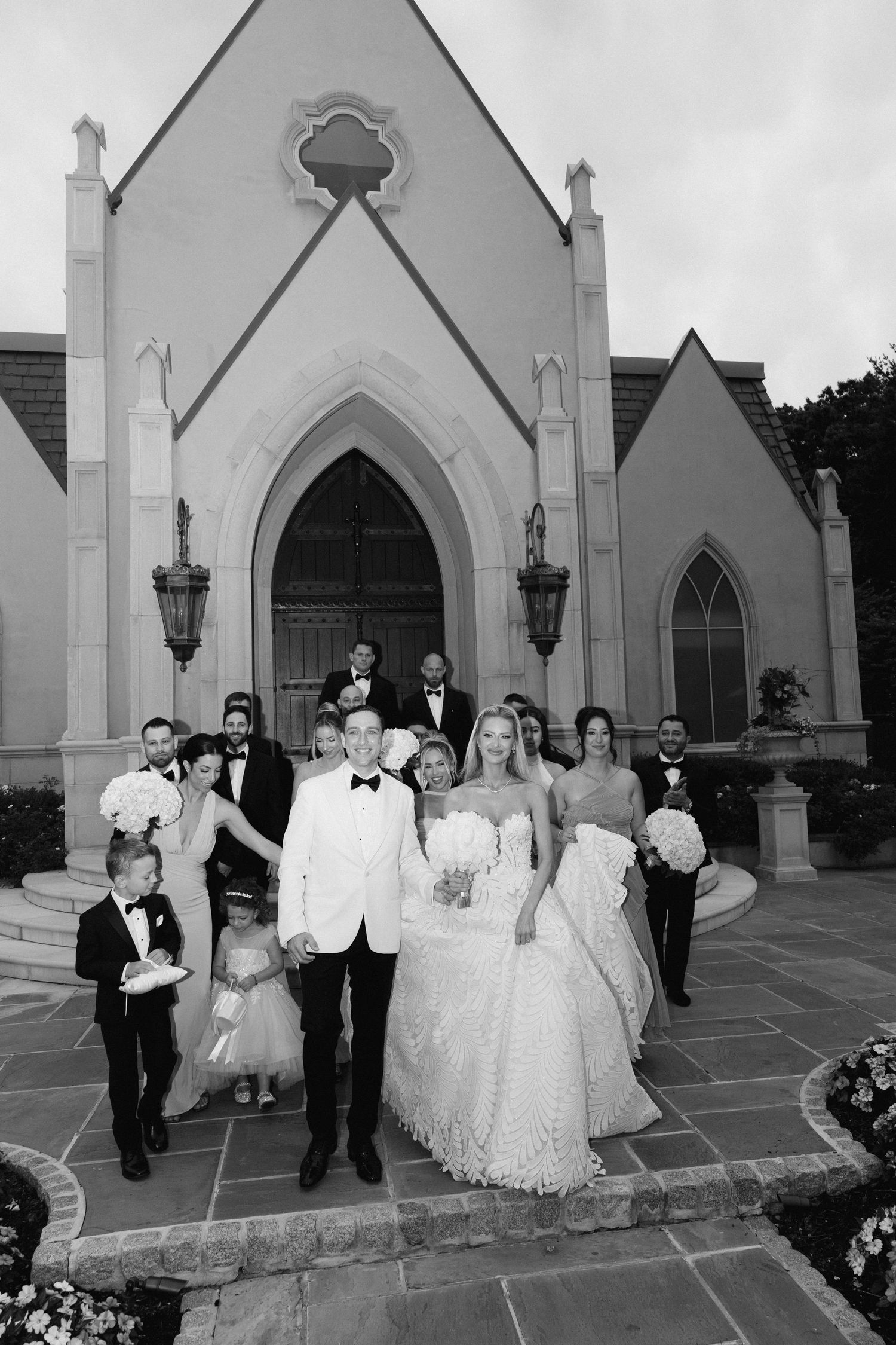 A wedding party stands on stone steps outside a Gothic-style church with arched doorways and lanterns.