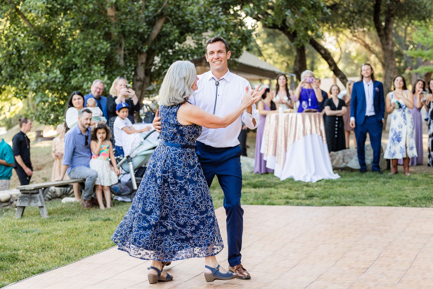 Couples dancing together at an outdoor wedding reception under trees with guests watching in the background.
