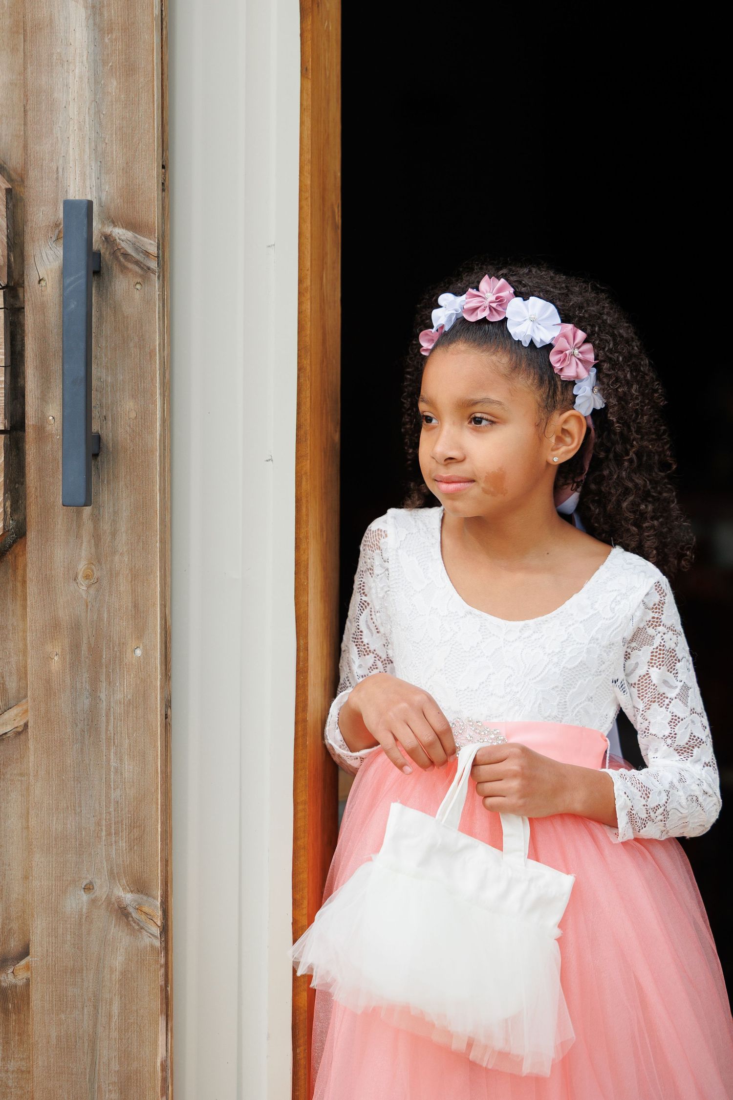 Young child in a white lace and pink tulle dress poses near a wooden doorframe with pink floral hair accessories.