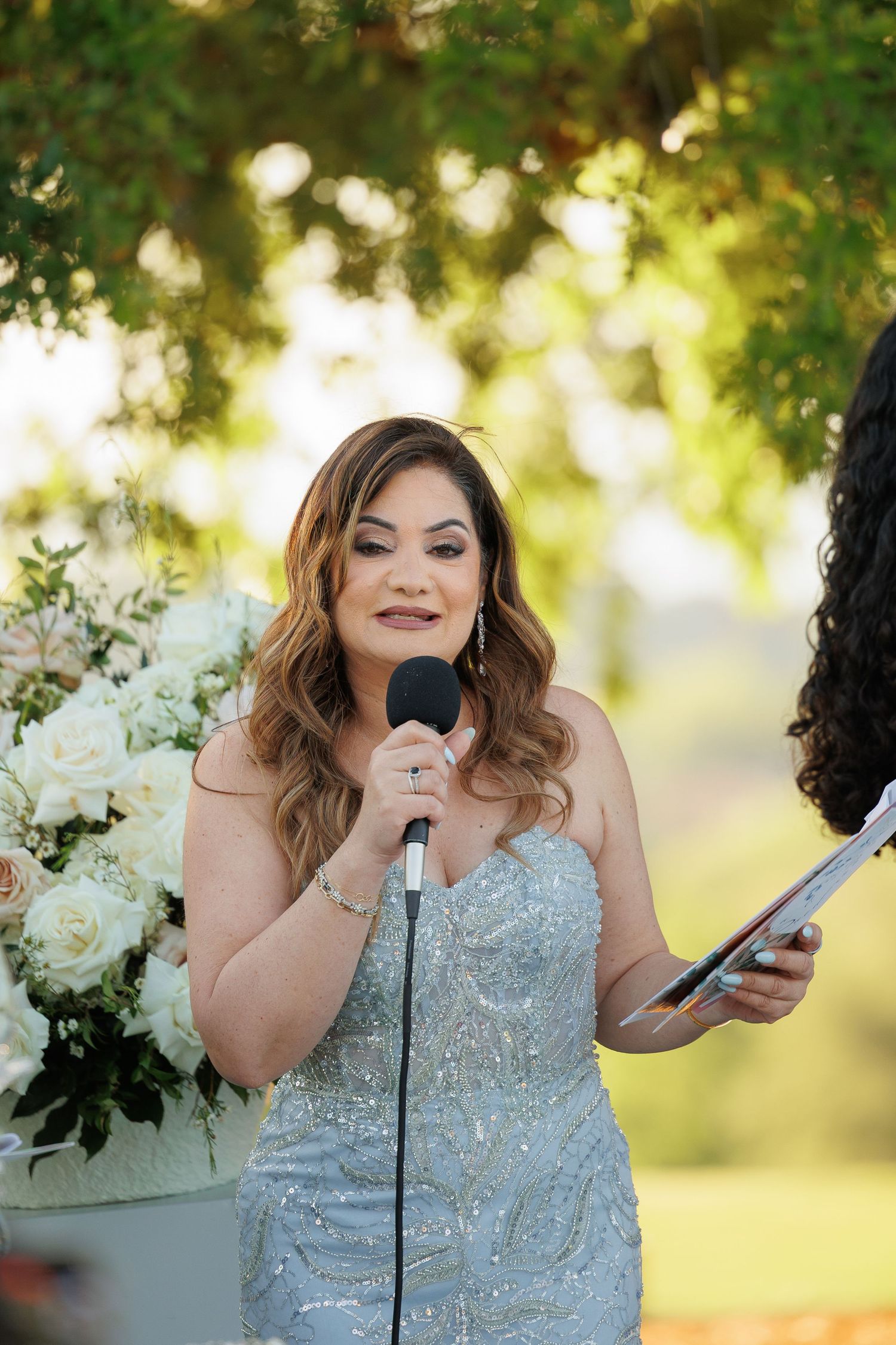 A person in a sparkly silver dress speaks into a microphone during an outdoor event with floral decorations.