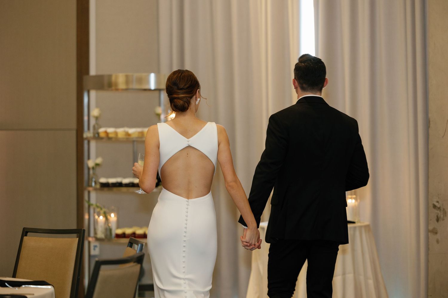 A wedding party walks down an elegant hallway with white curtains and golden lighting.