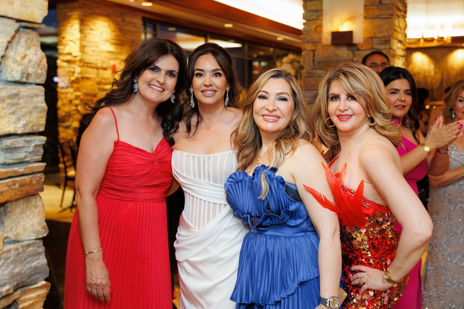A group of people in colorful formal attire pose together at an indoor event with stone walls and warm lighting.